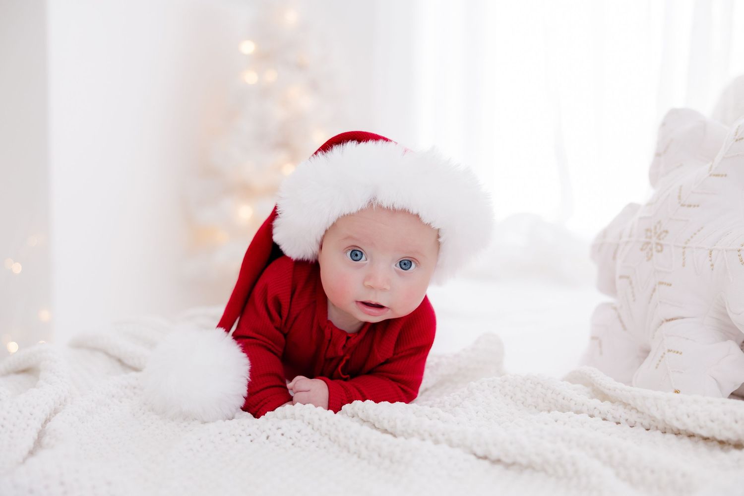 Baby wearing red Santa outfit crawling on white blanket with Christmas lights in background.