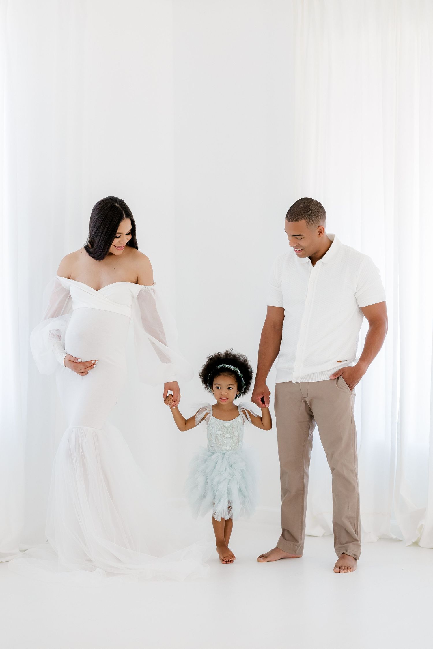 Family maternity photoshoot in all white with toddler daughter dancing between parents.