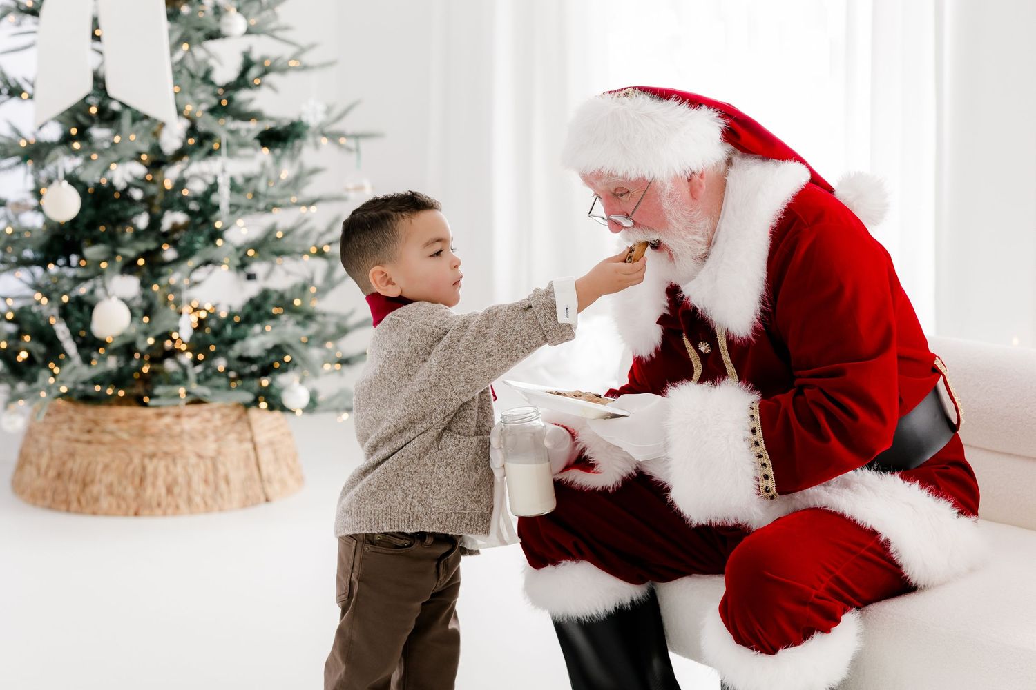 A young boy feeds a cookie to Santa Claus beside a decorated Christmas tree in a bright white room.