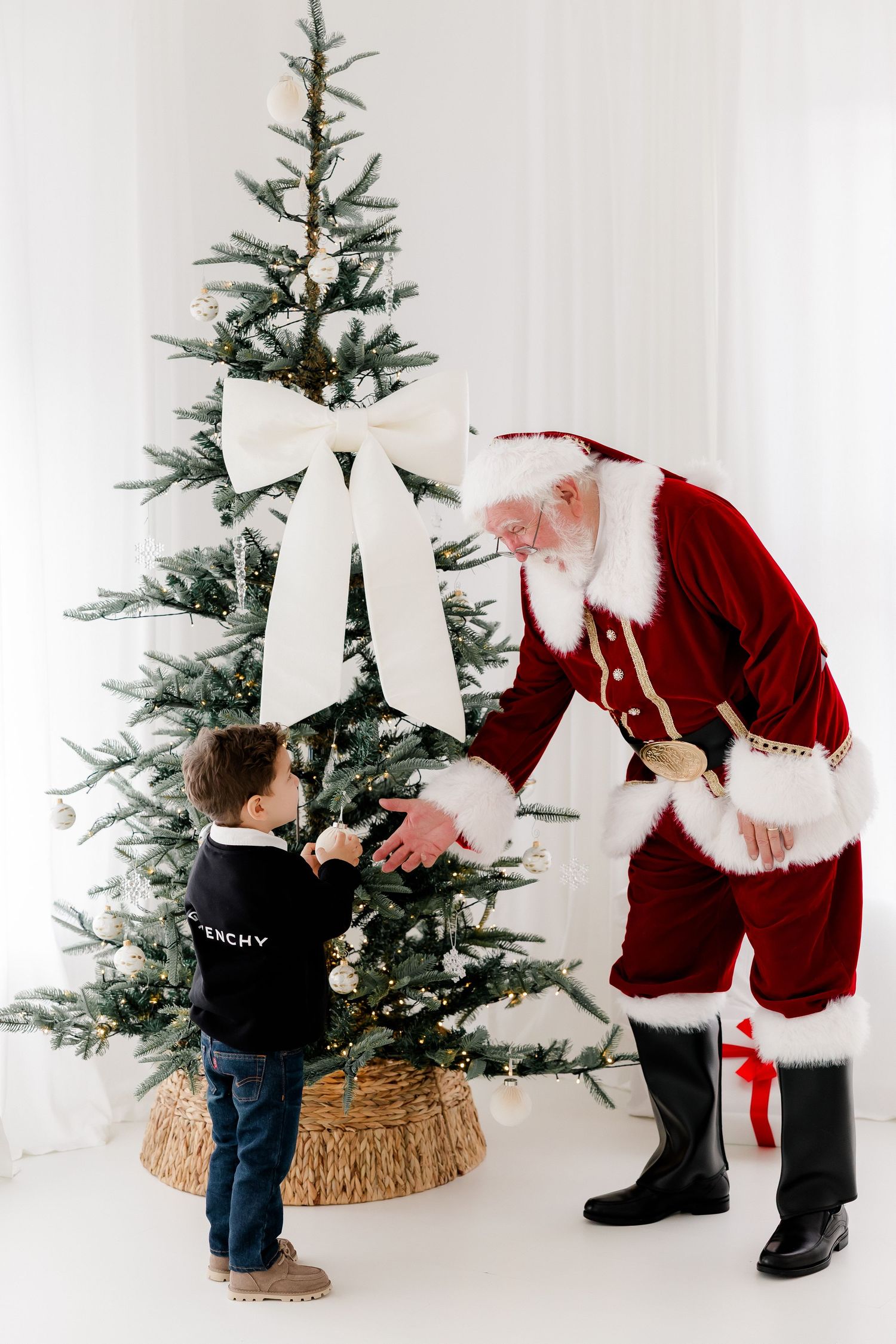 Child meeting Santa Claus by a decorated Christmas tree with a large white bow in a bright white room.