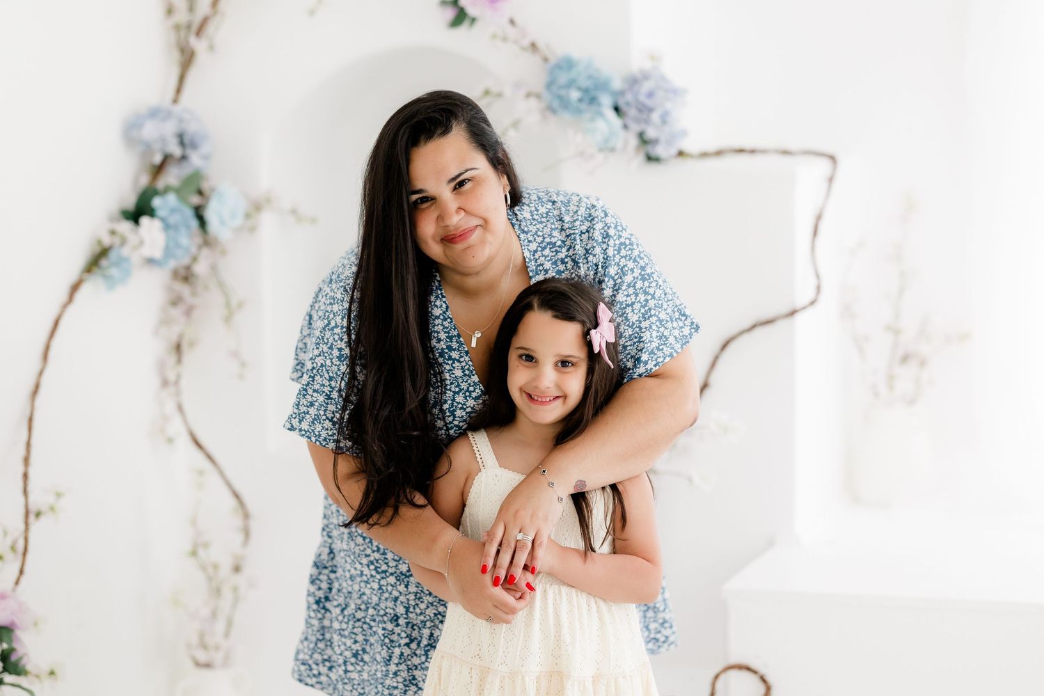 A parent and child embrace while wearing a blue floral dress and white dress against a light backdrop.