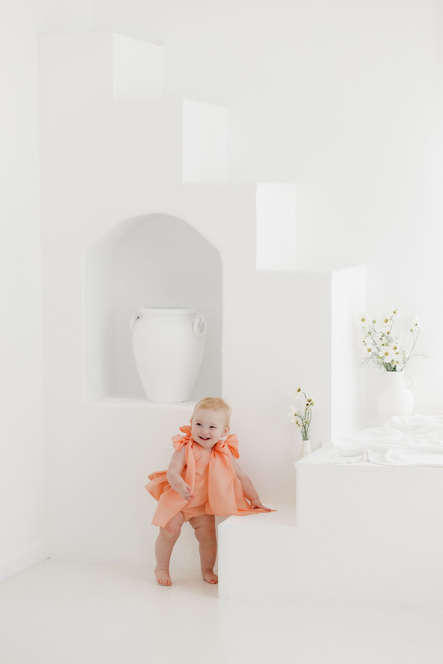 A small child wearing a peach-colored outfit stands in a minimalist white room with built-in wall niches and delicate flowers.