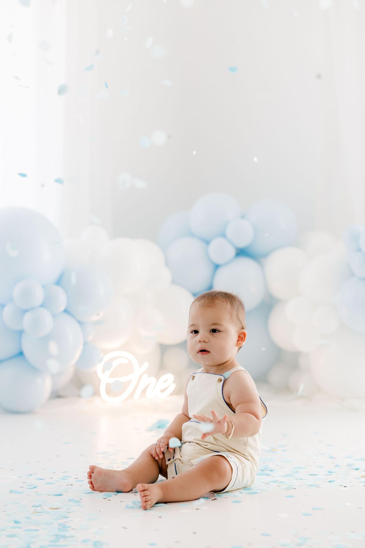 Baby in white onesie sitting against dreamy light blue balloon backdrop for milestone photo session.