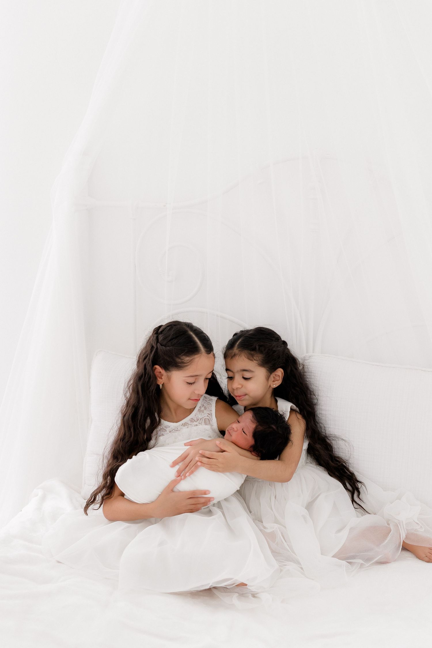 Two young girls in white dresses sitting together on white bedding while holding and looking at a baby.
