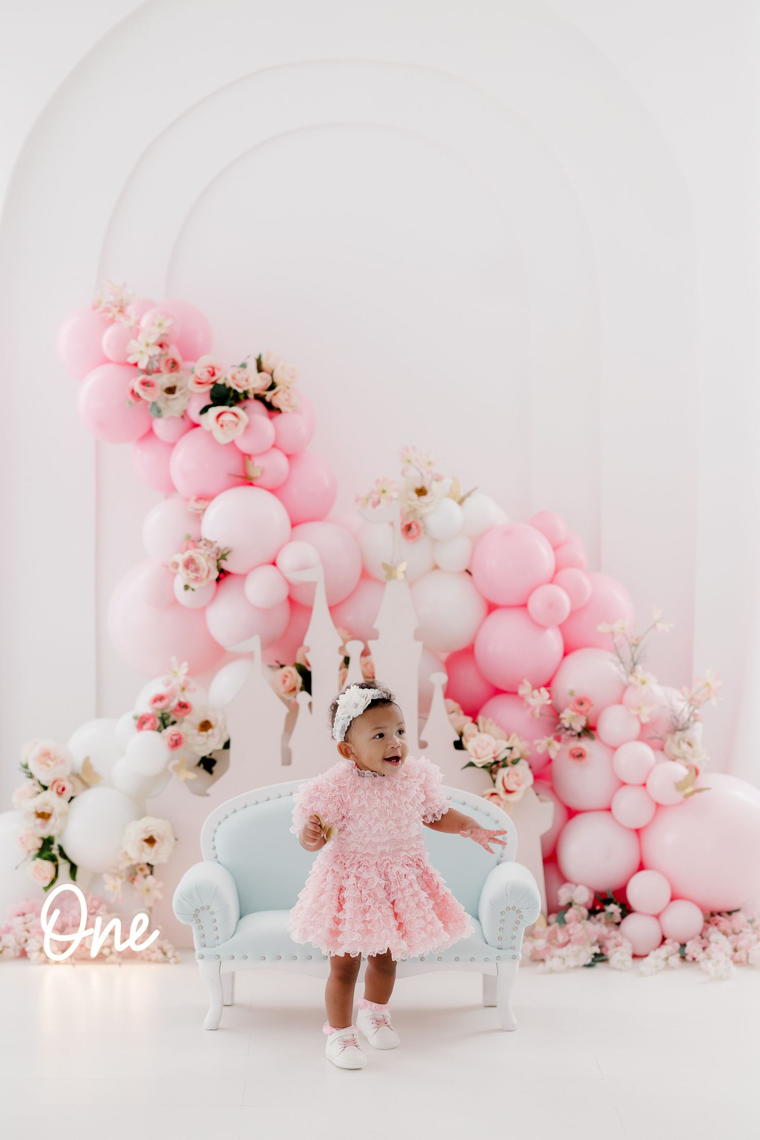 Elegant pink and white balloon display with floral accents and a vintage-style chair set against white backdrop.