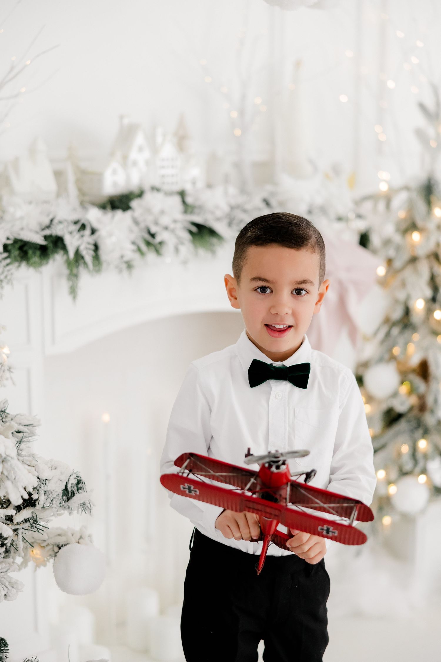 Young boy in formal attire holding red toy airplane in white Christmas setting.nj holiday mini sessions