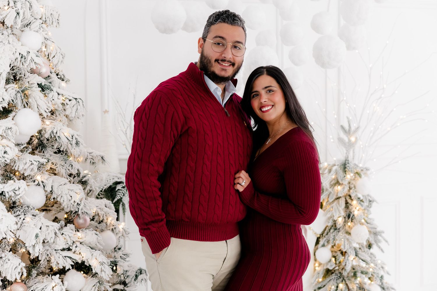 Couple in coordinating burgundy sweaters posing by white Christmas tree in studio setting.