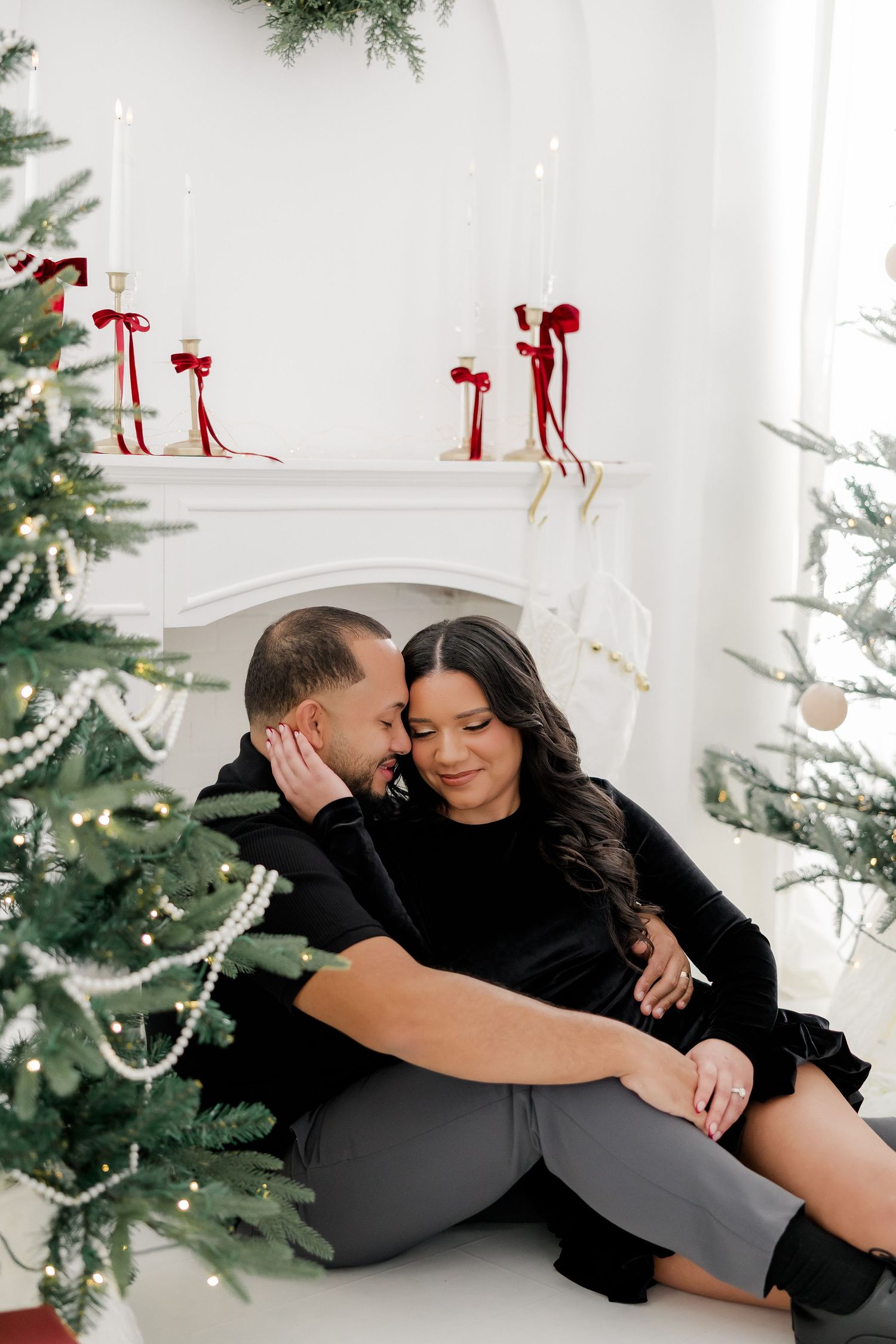 Romantic couple in black outfits sitting together by a white fireplace with Christmas trees and red ribbon candles.