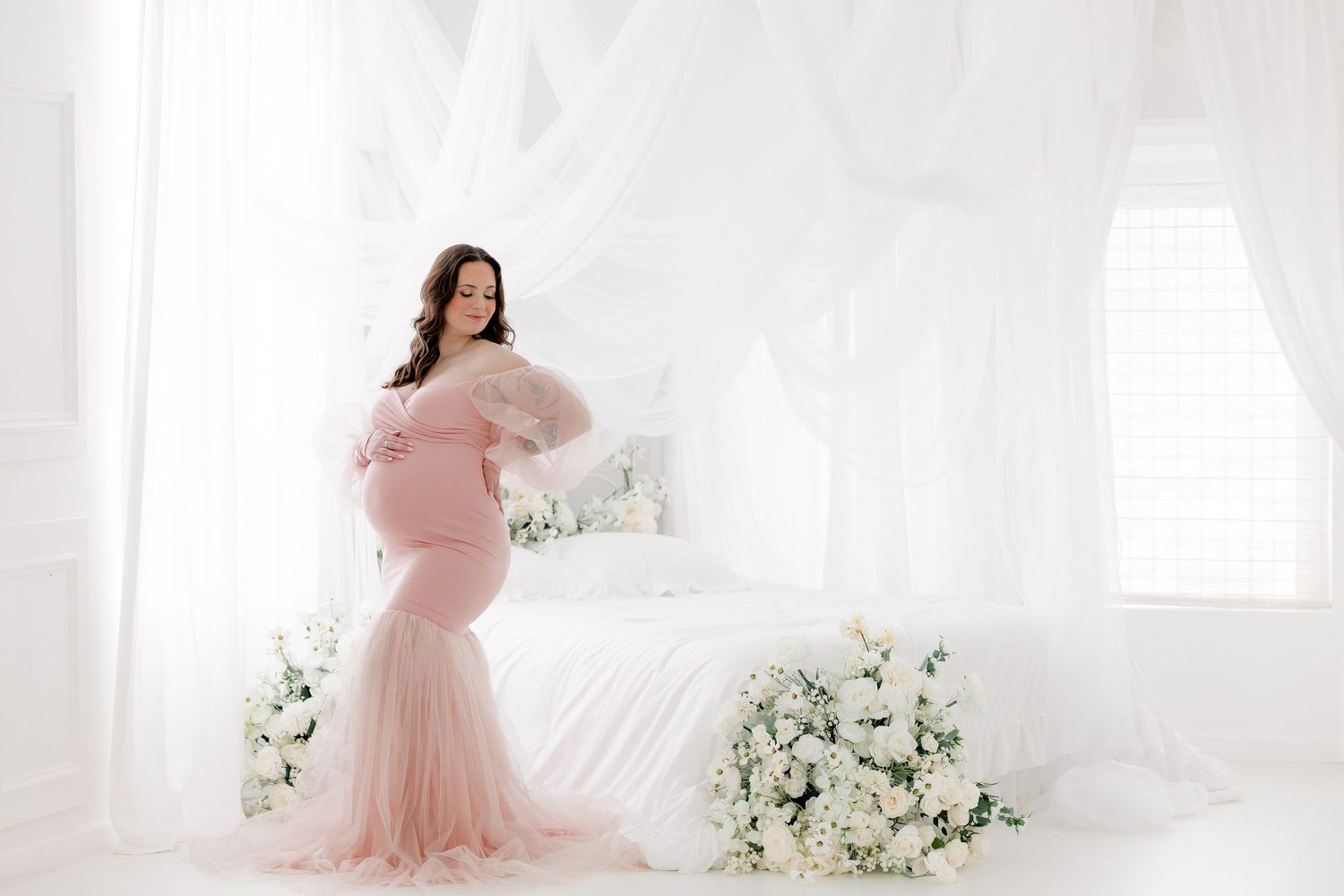 A dreamy maternity portrait in a light-filled white room with blush pink tulle gown and white floral arrangements.