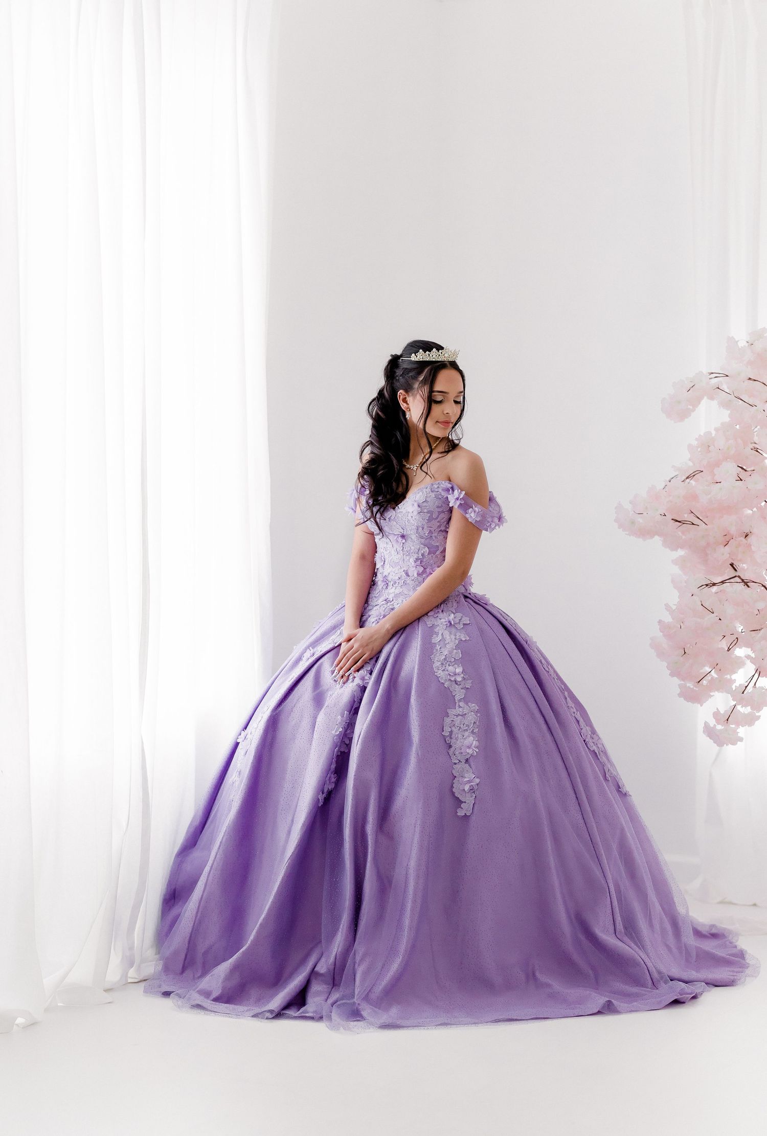 A lavender ball gown with off-shoulder details and lace appliques photographed in a white studio setting.