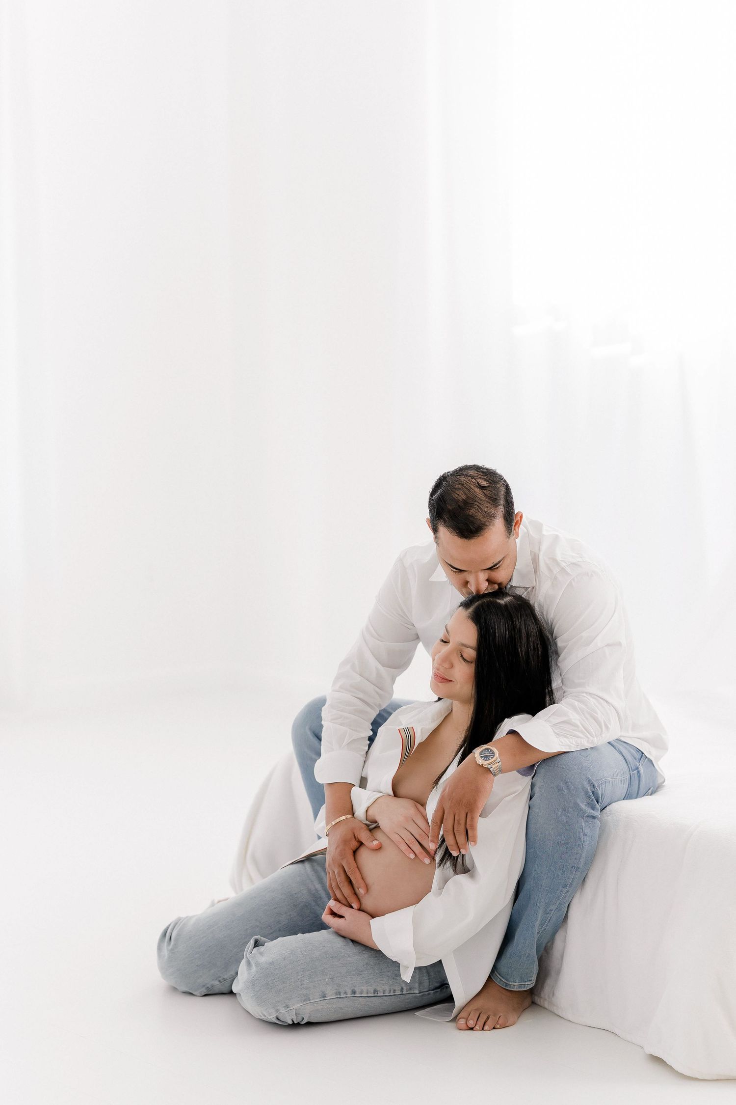 Intimate couple wearing white and denim embrace in front of white backdrop.