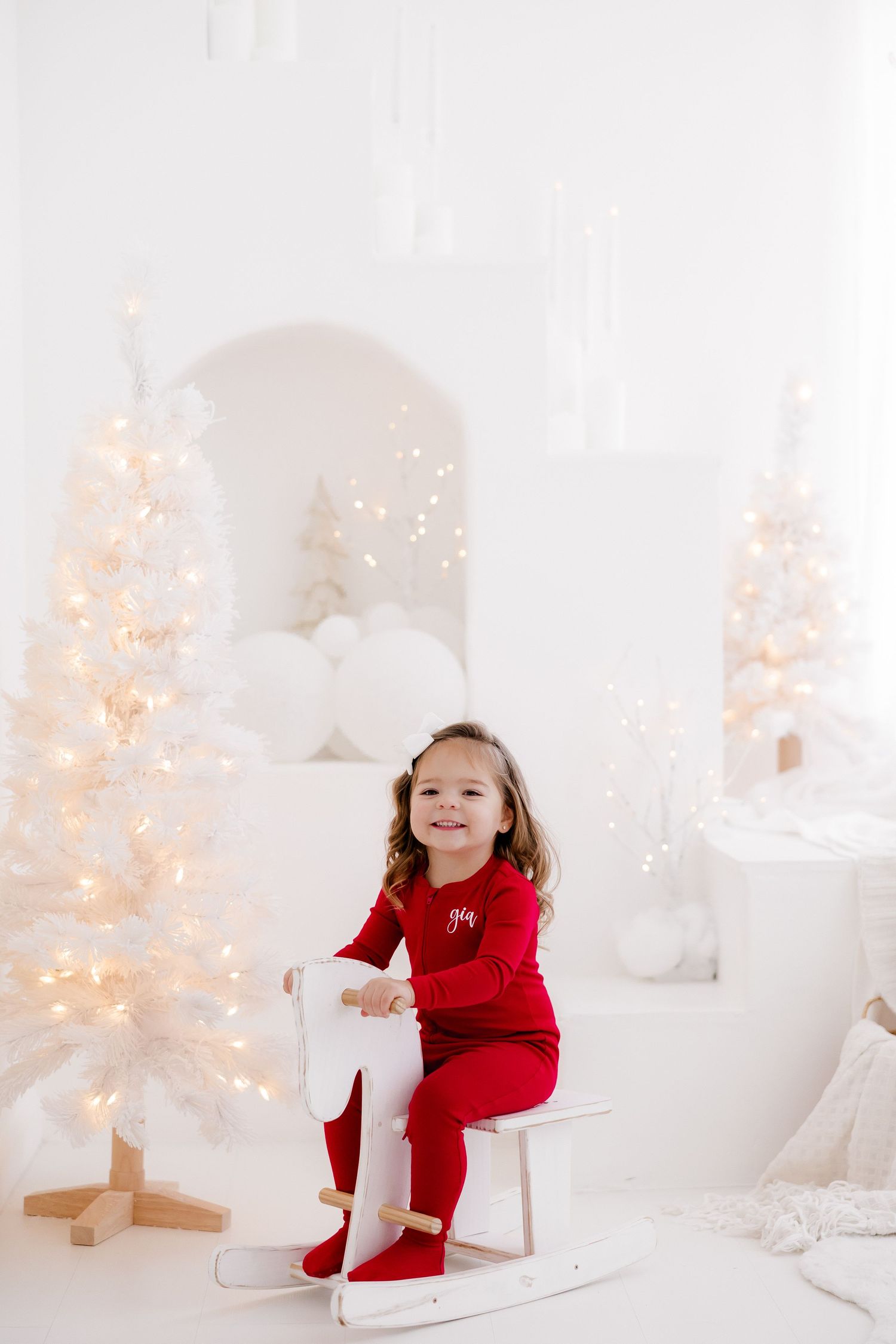 Child in red outfit sitting on rocking chair surrounded by white lit Christmas trees.