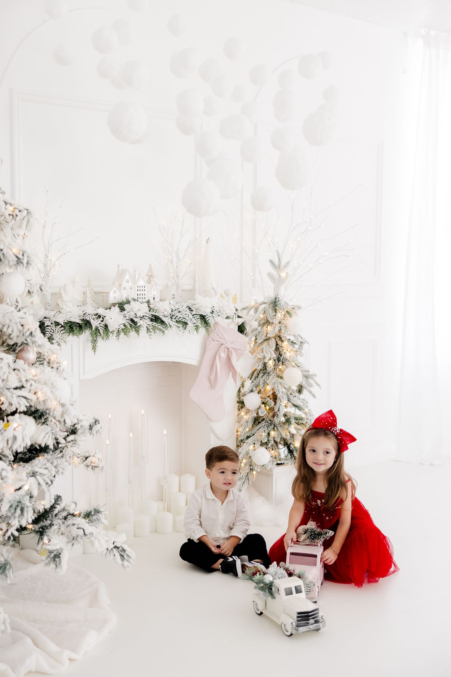 Children in holiday outfits play by decorated Christmas trees in an all-white studio setting.