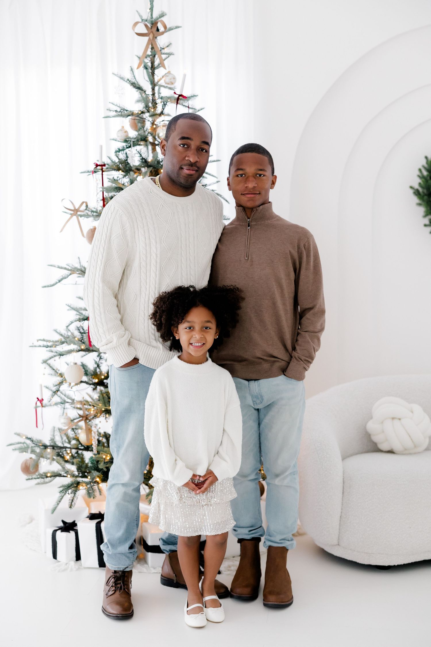 Family in coordinated casual outfits posing near a decorated Christmas tree in a white studio.