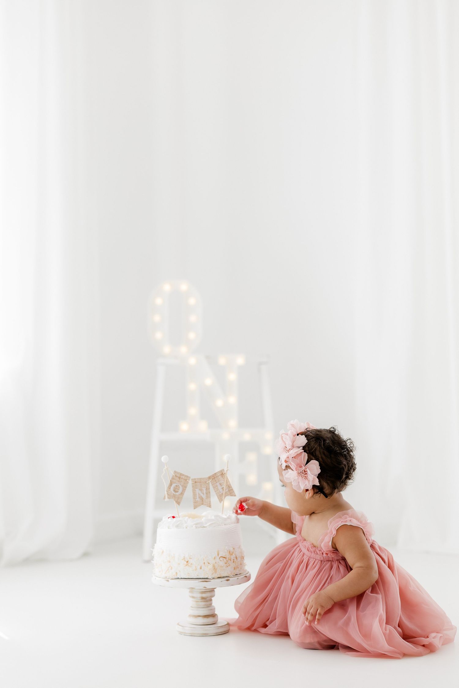 A figure in a flowing pink dress reclines on white floor near an elegant white cake with decorative bunting.