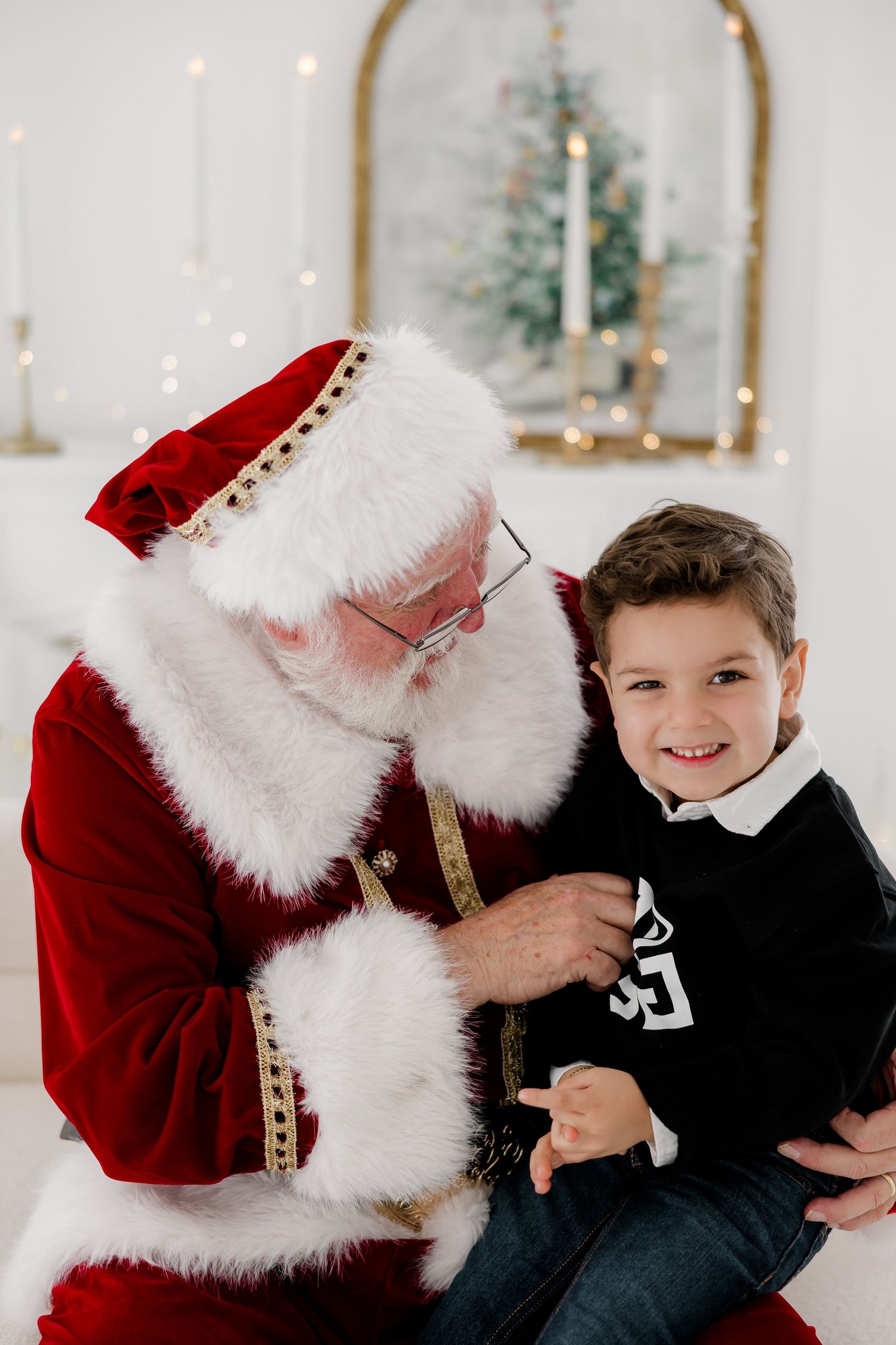 A smiling young boy sits on Santa Claus's lap during a cheerful Christmas portrait session with holiday decorations behind them.
