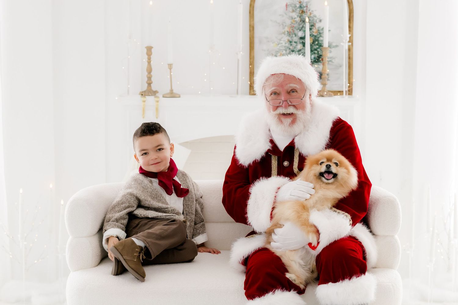 A young boy and Santa Claus sit on a white chair holding a Pomeranian dog in a bright holiday photo setting.
