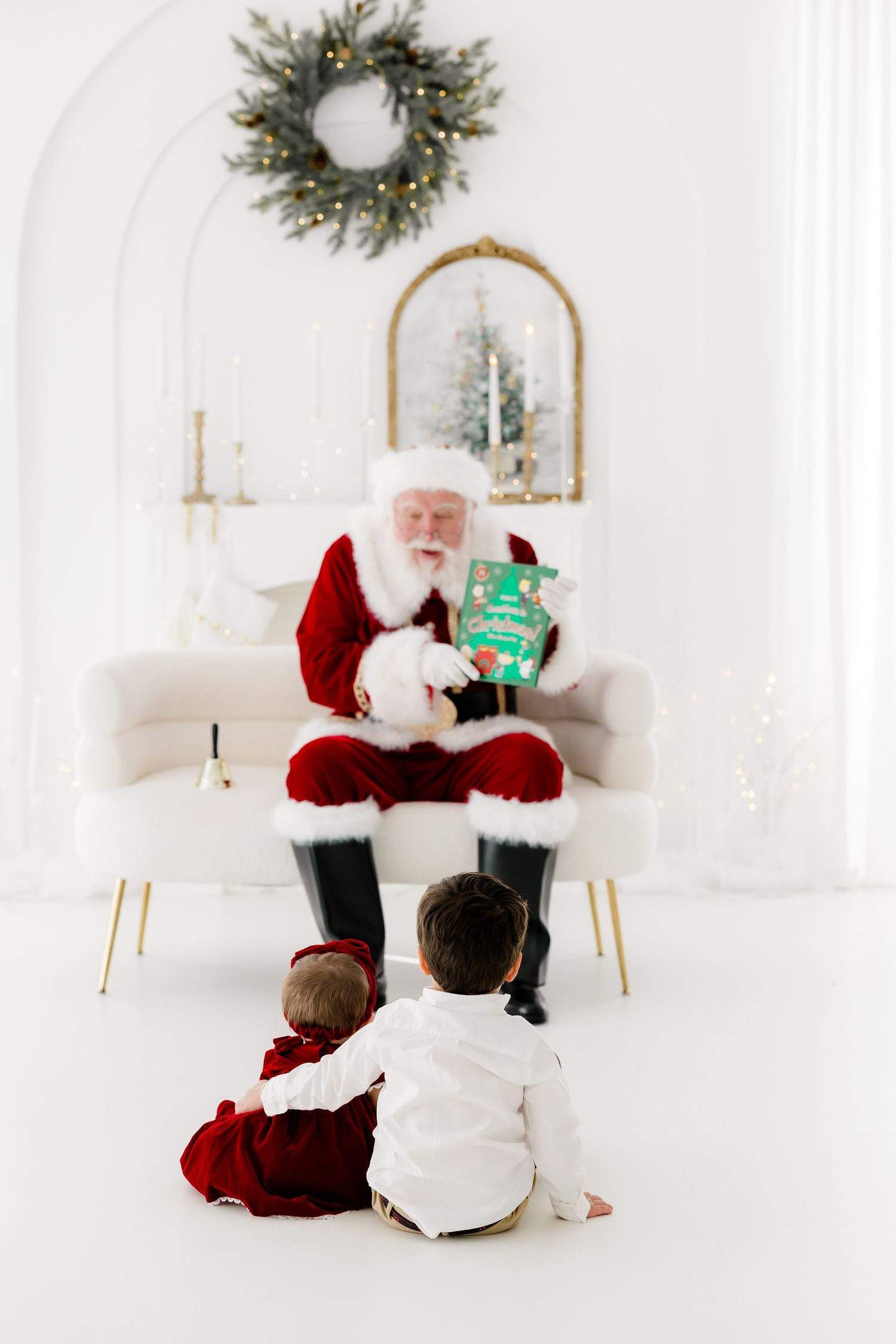 Two toddlers sit on floor facing Santa Claus holding a gift in a bright white Christmas decorated room.