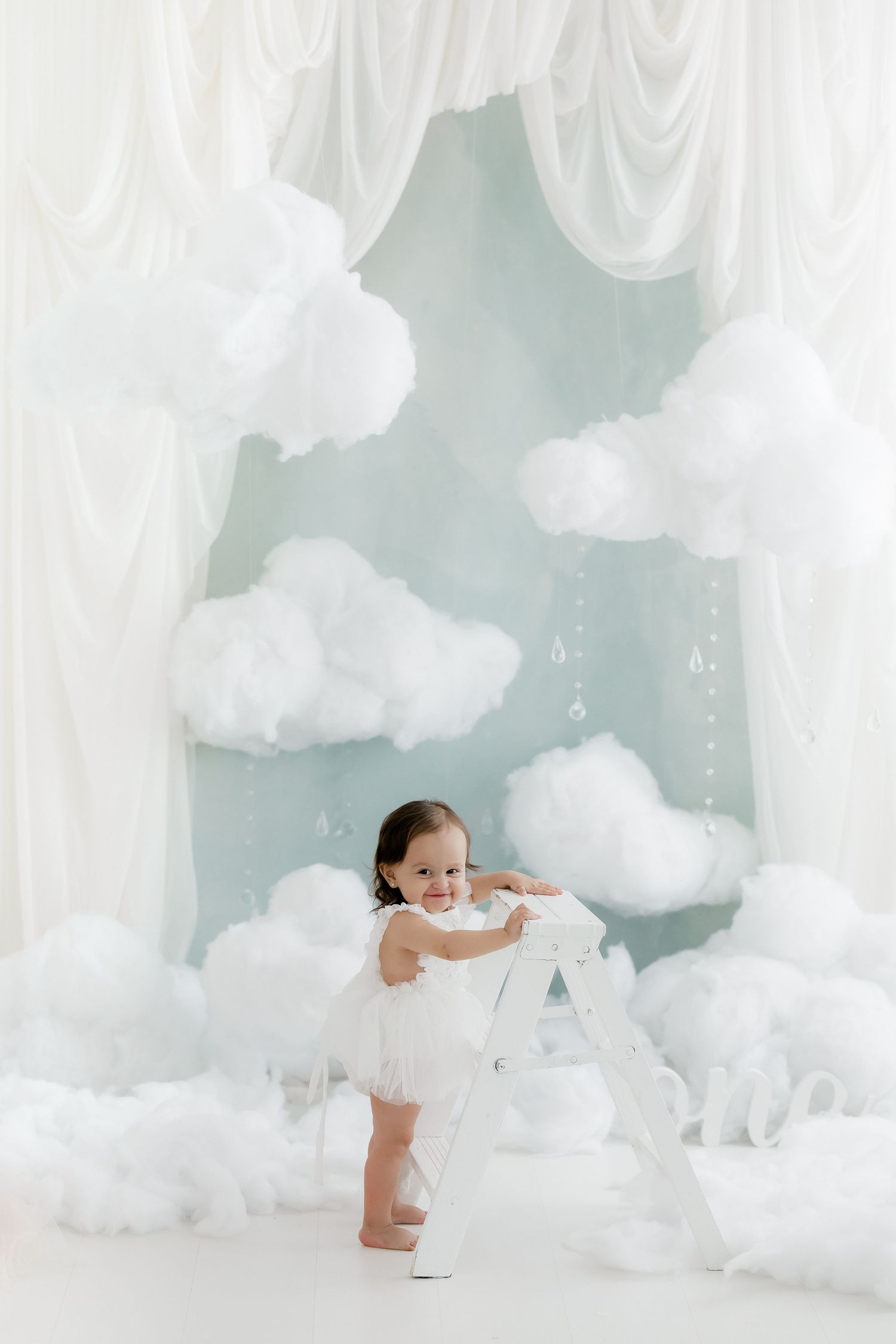 Toddler in white dress standing by letter A prop in dreamy cloud-themed photo studio backdrop.
