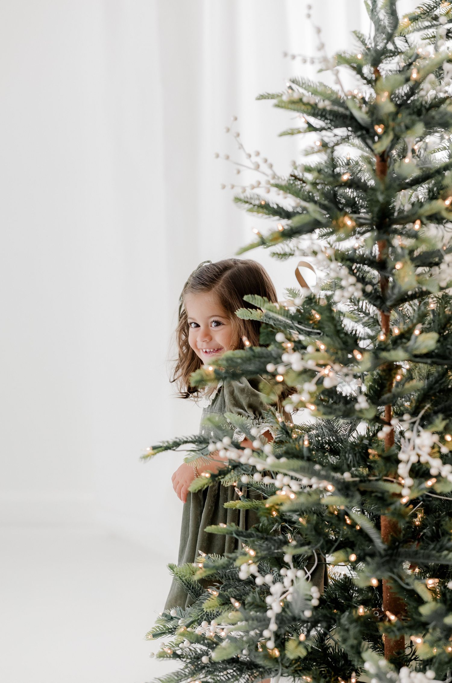A smiling young girl in a green dress peeking out from behind a decorated Christmas tree with lights.
