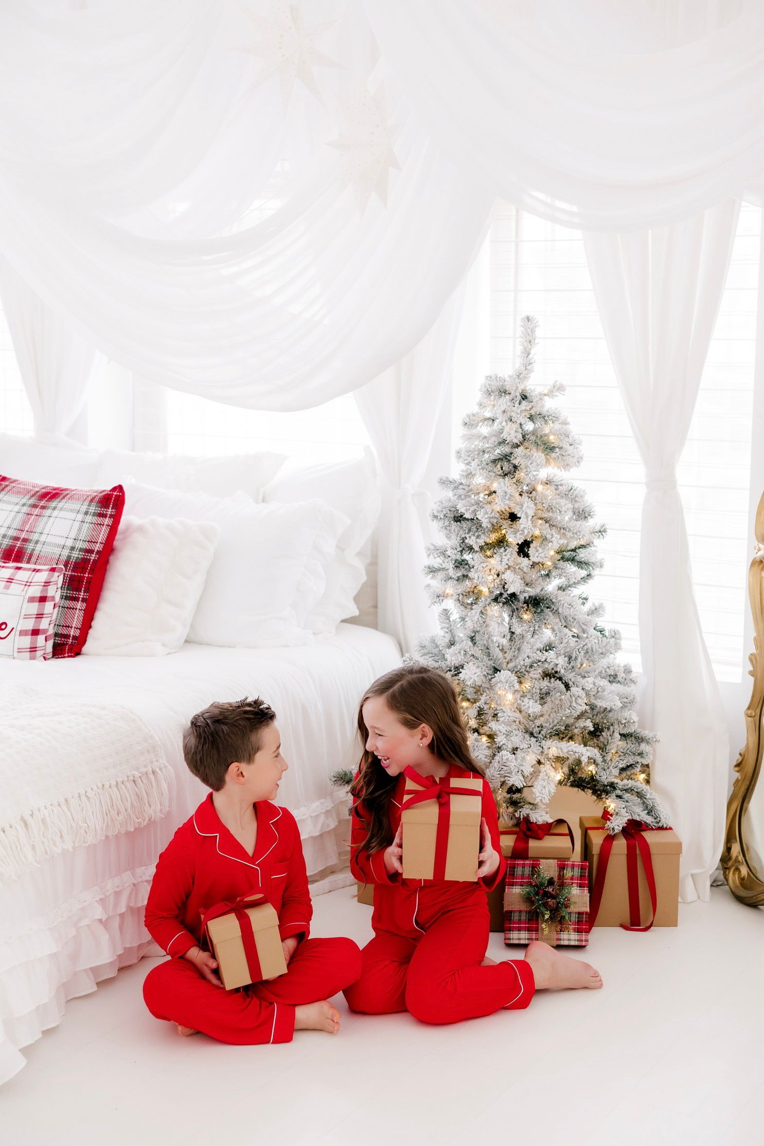 Two children in red pajamas exchange Christmas gifts beside a flocked tree in a white decorated bedroom.