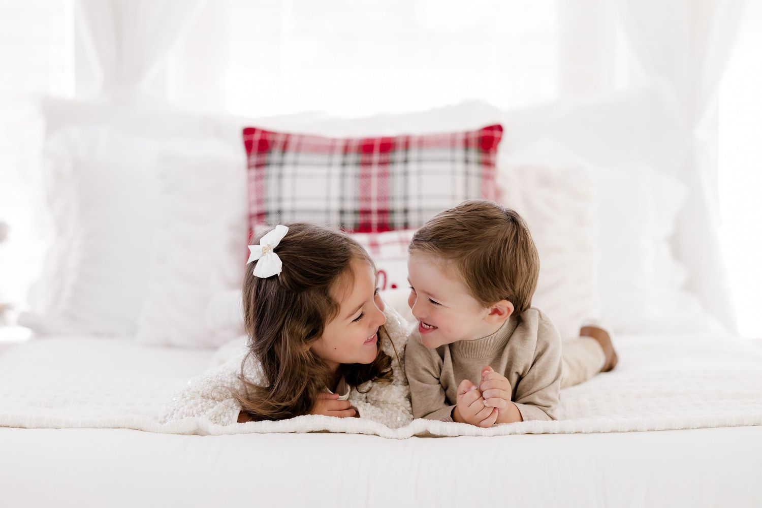 Two young children lying on a white bed, smiling at each other, with a plaid holiday pillow in the background.