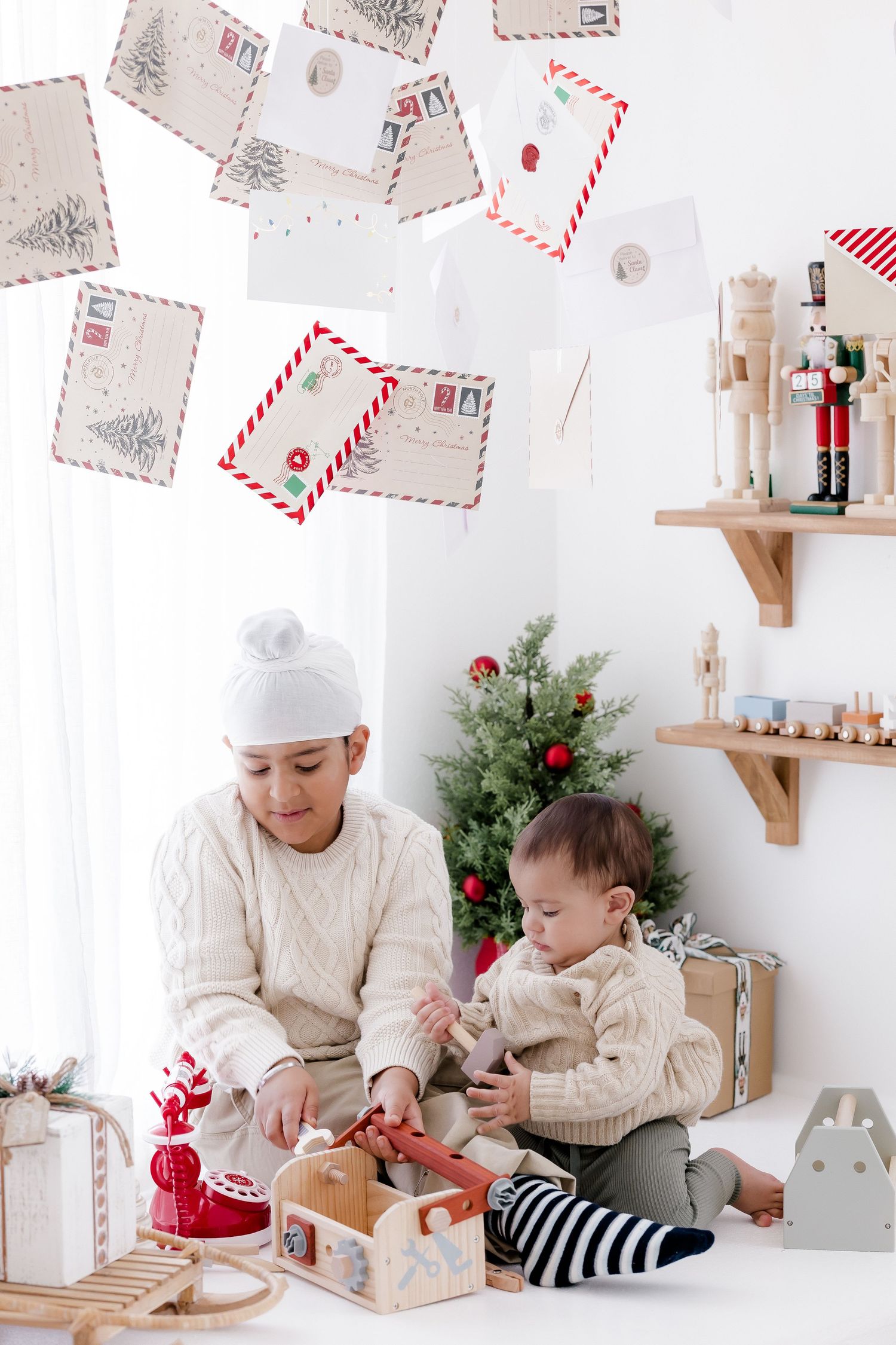 Two children playing with wooden Christmas toys in a festive white room with holiday cards, a tree, and nutcracker decorations.