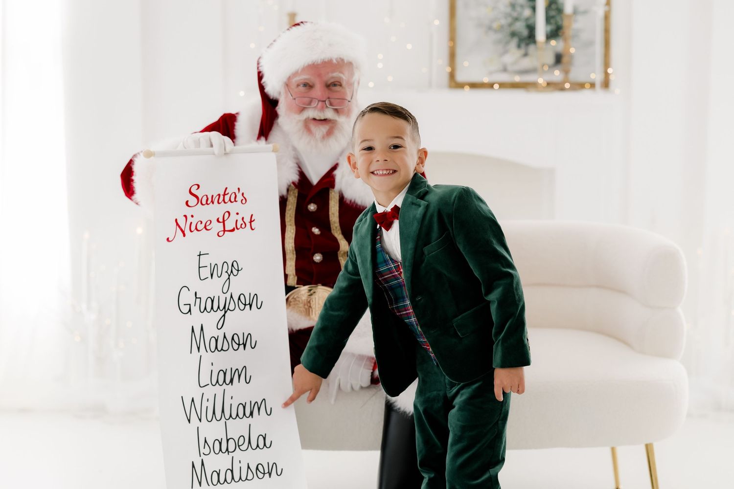Young boy in green velvet suit poses with Santa Claus holding a Nice List scroll in a bright white holiday studio.