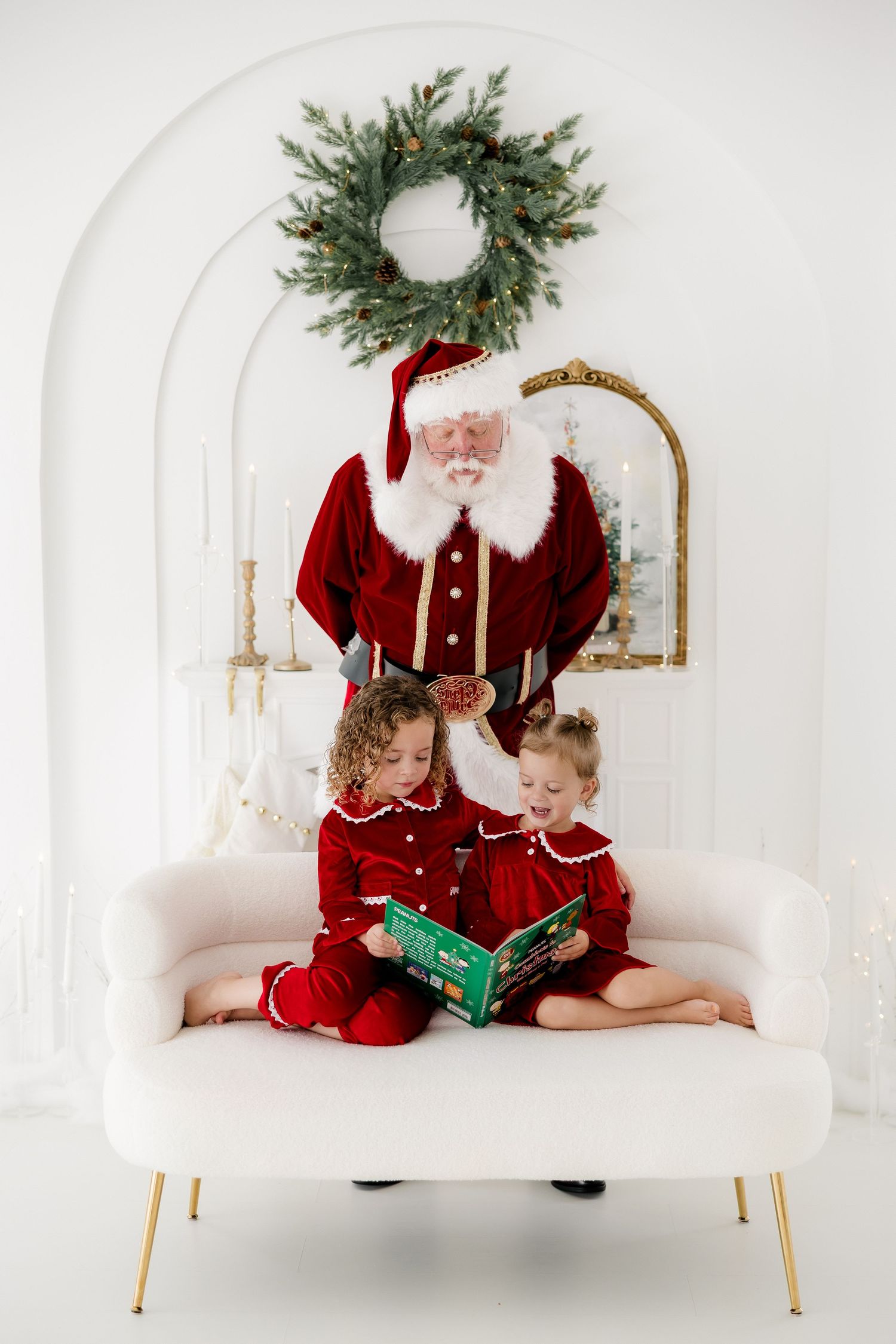 Two children in red outfits read a book with Santa Claus on a pink sofa in a festive white room with a Christmas wreath.