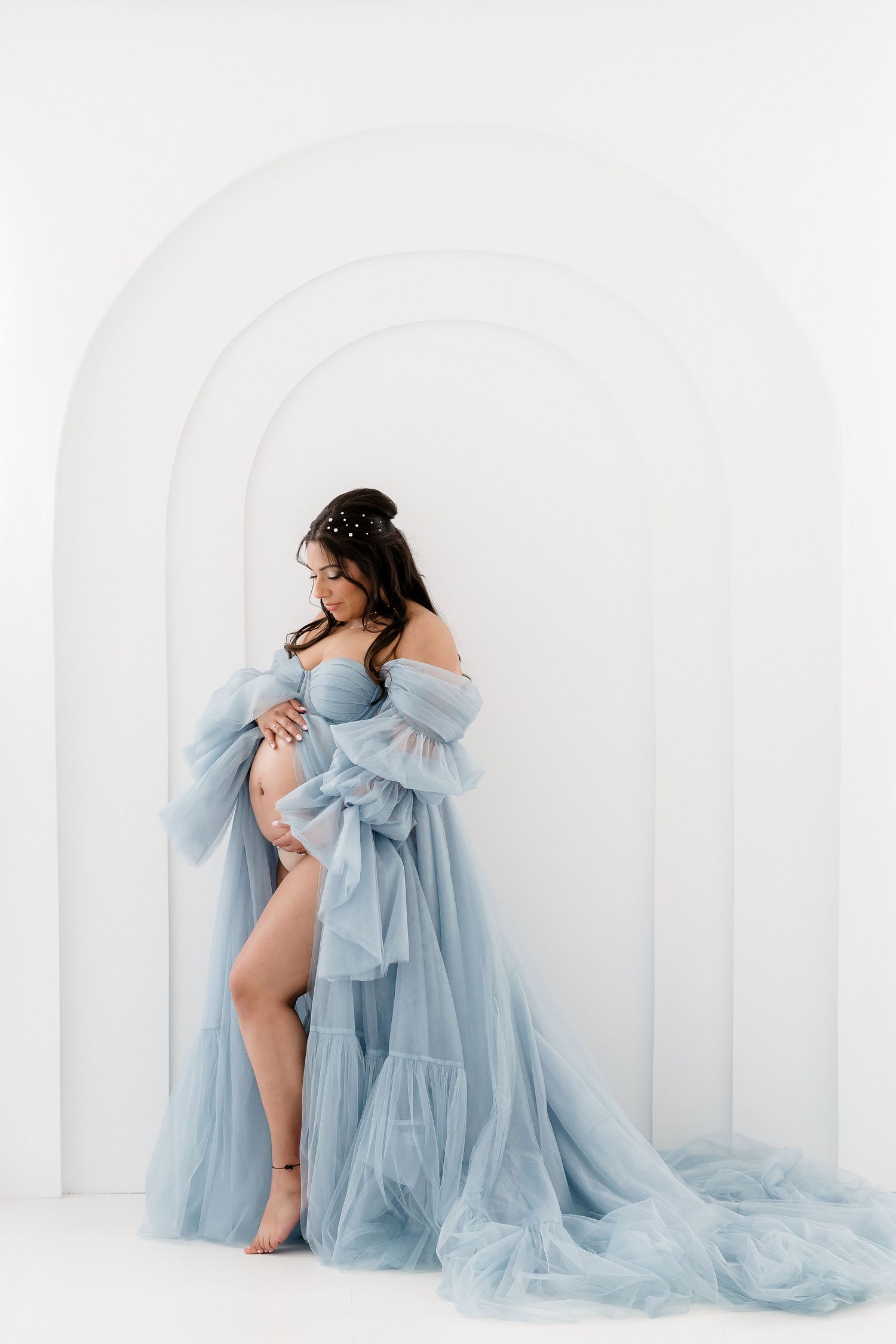 Ethereal maternity photoshoot featuring flowing light blue tulle gown against white backdrop in artistic poses.