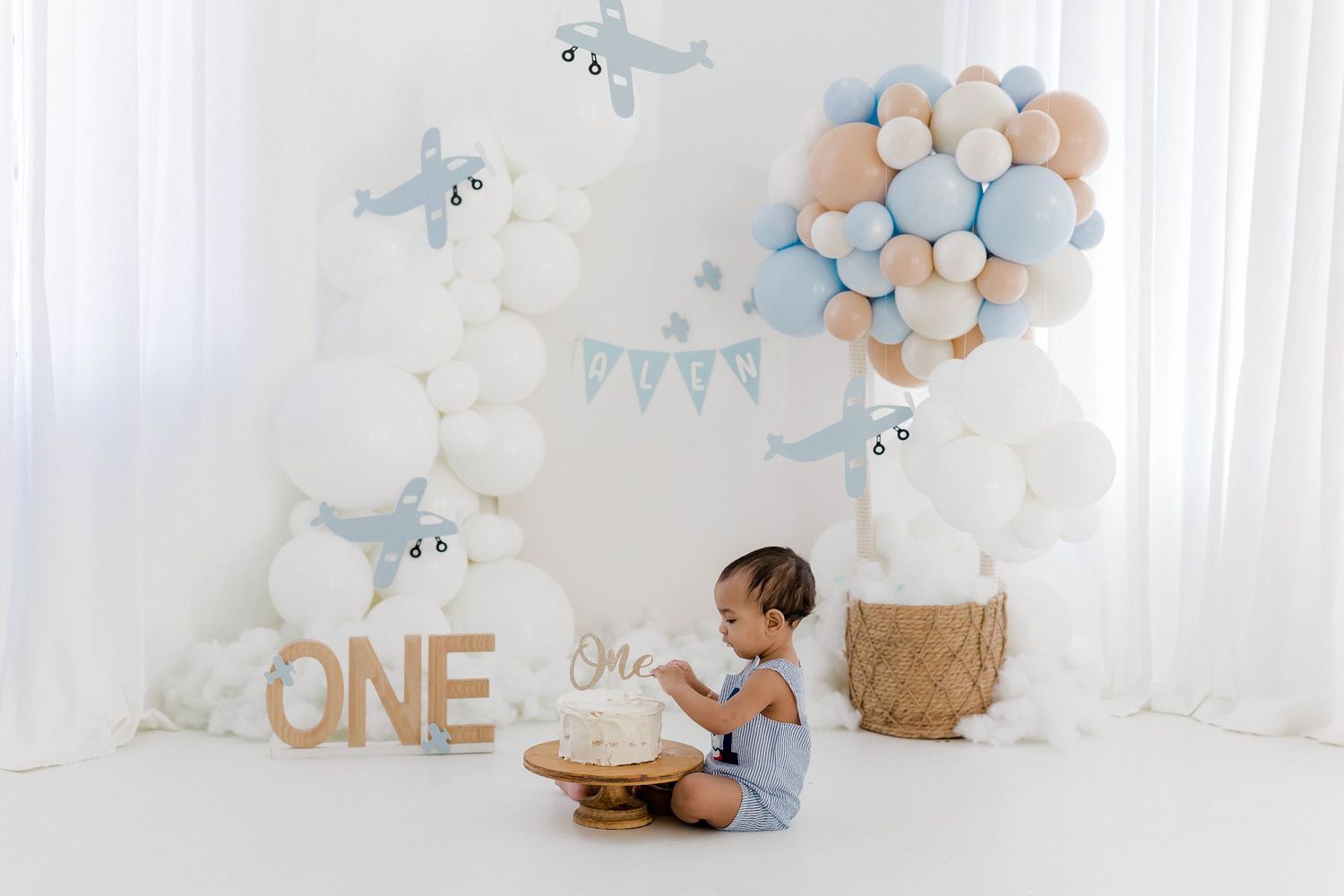 A pastel blue and gold first birthday party setup with balloons, banners, and decorative hot air balloon baskets.