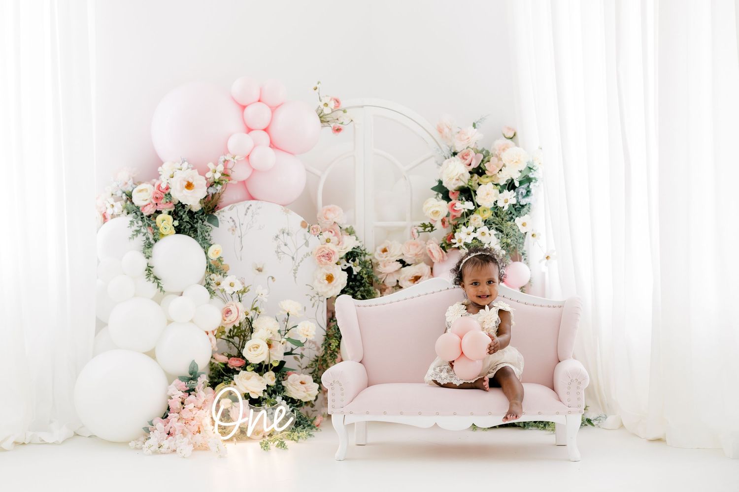 Elegant pink and white floral decorations with balloons create a romantic setting with plush cushions and soft lighting.