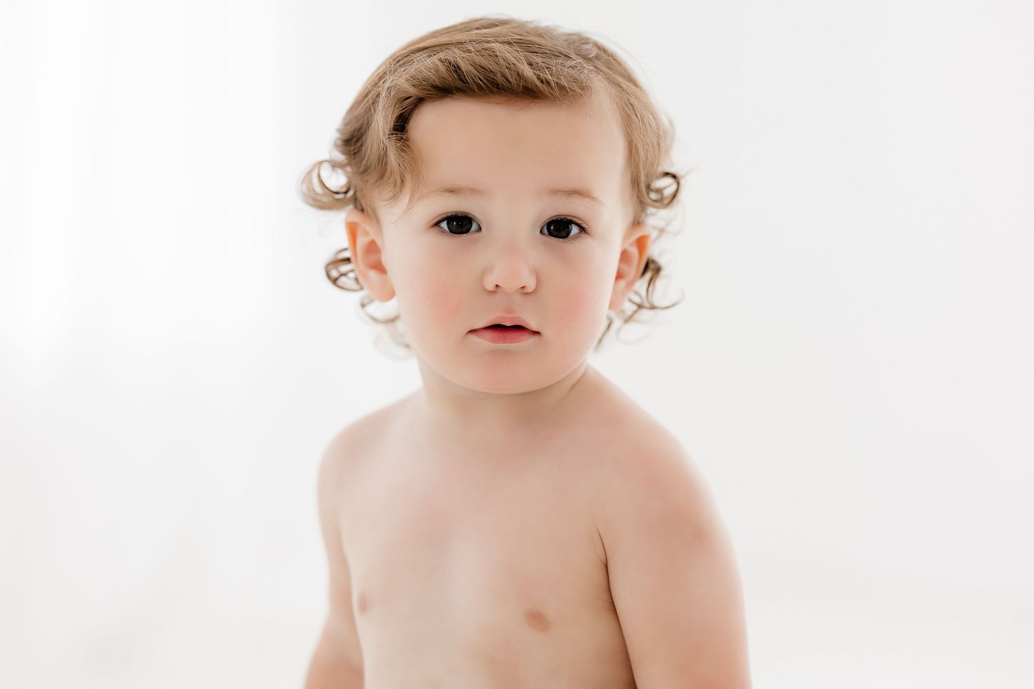 A small child with curly blonde hair sits against a white background in a minimalist portrait setting.