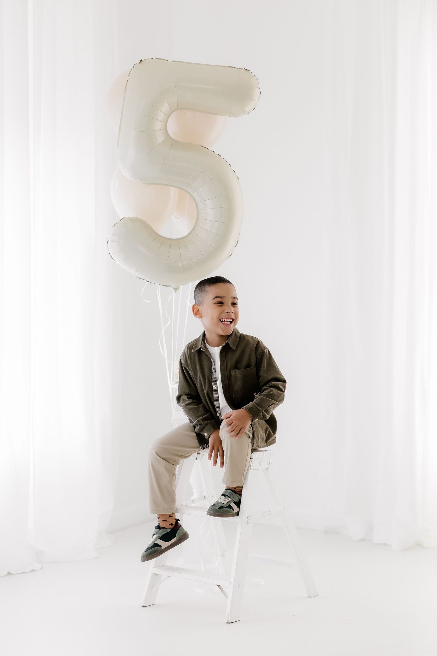 A young child sits on a white stool with a large number 5 balloon floating above in a minimalist white setting.