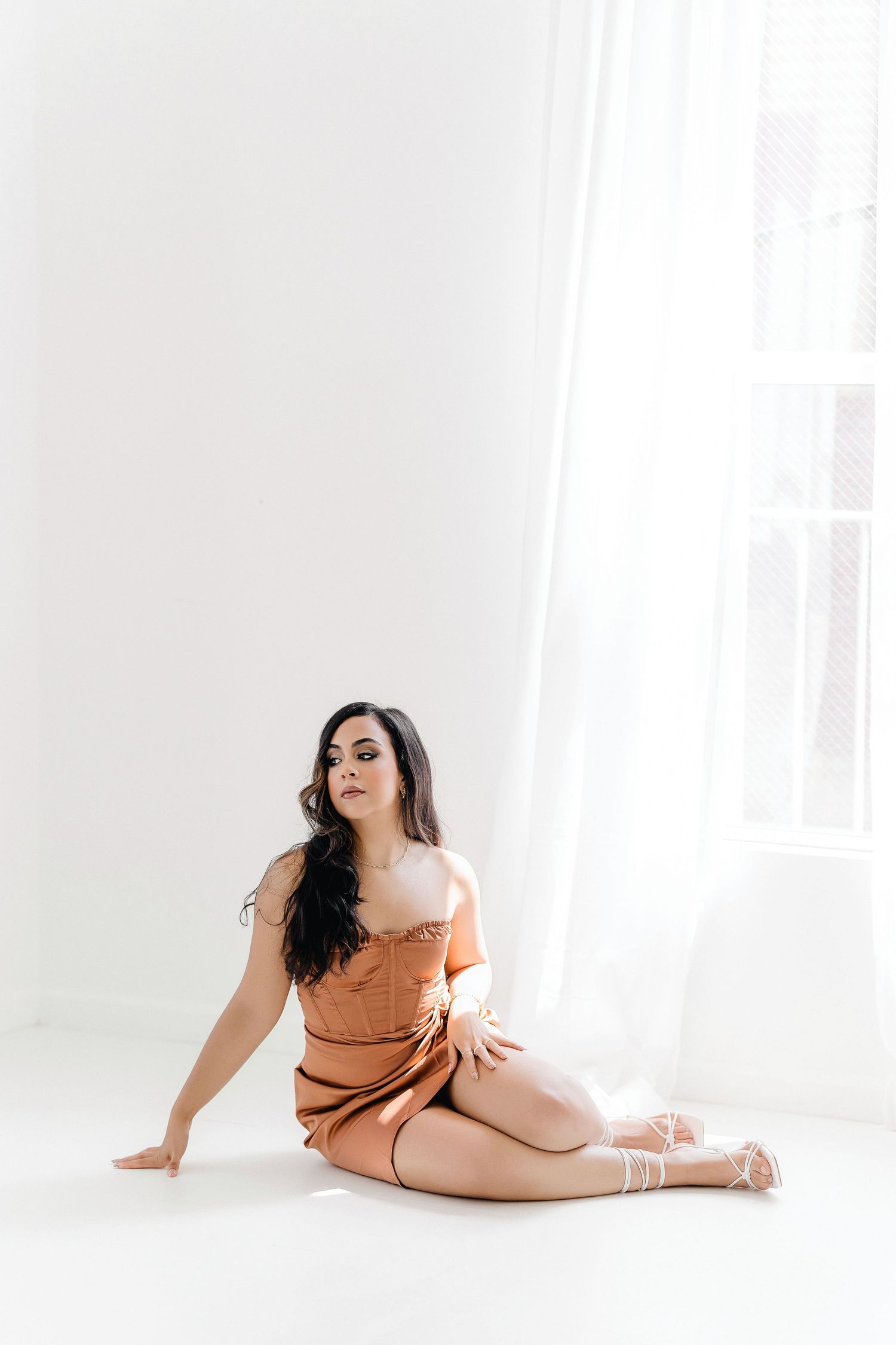Woman in orange bodysuit poses gracefully on white floor in bright minimalist studio setting.