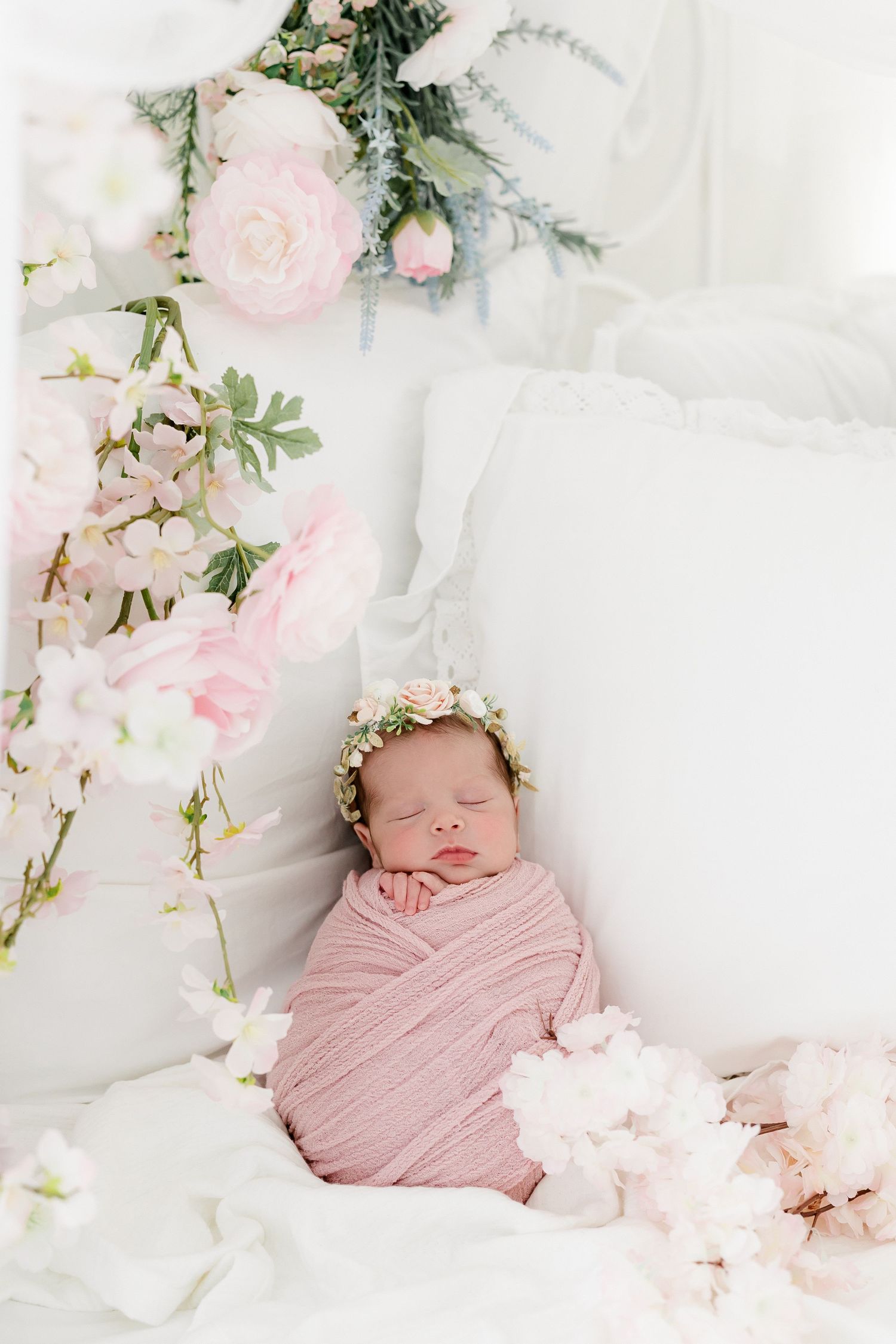 Newborn wrapped in soft pink fabric surrounded by delicate cherry blossoms and roses on white bedding.