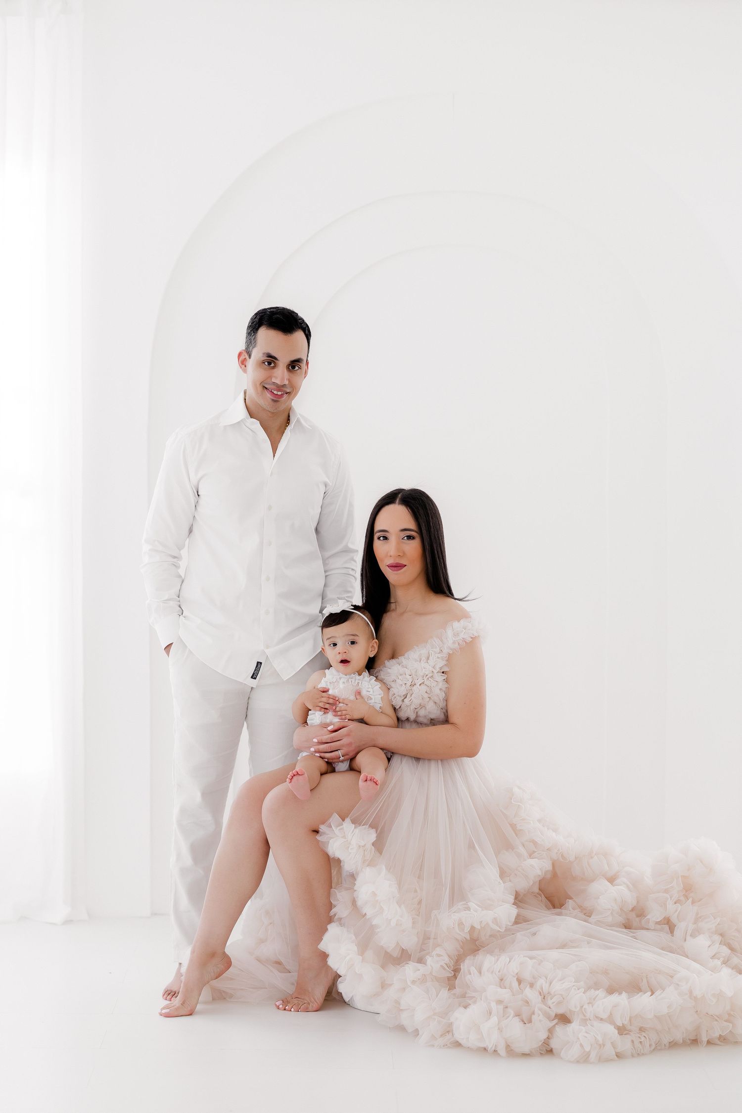 Elegant family portrait in all white against white background with flowing tulle gown and baby in formal attire.