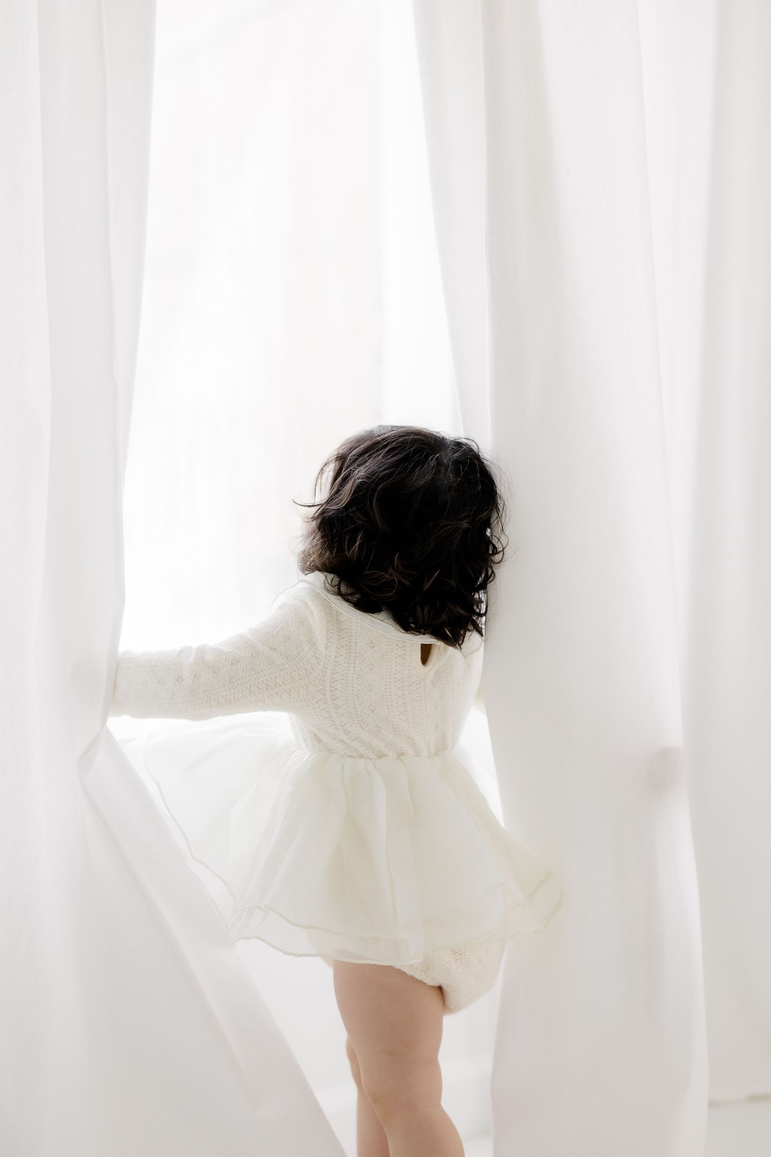 A young child in a white dress and sweater reaches for sheer white curtains in a bright, airy room.