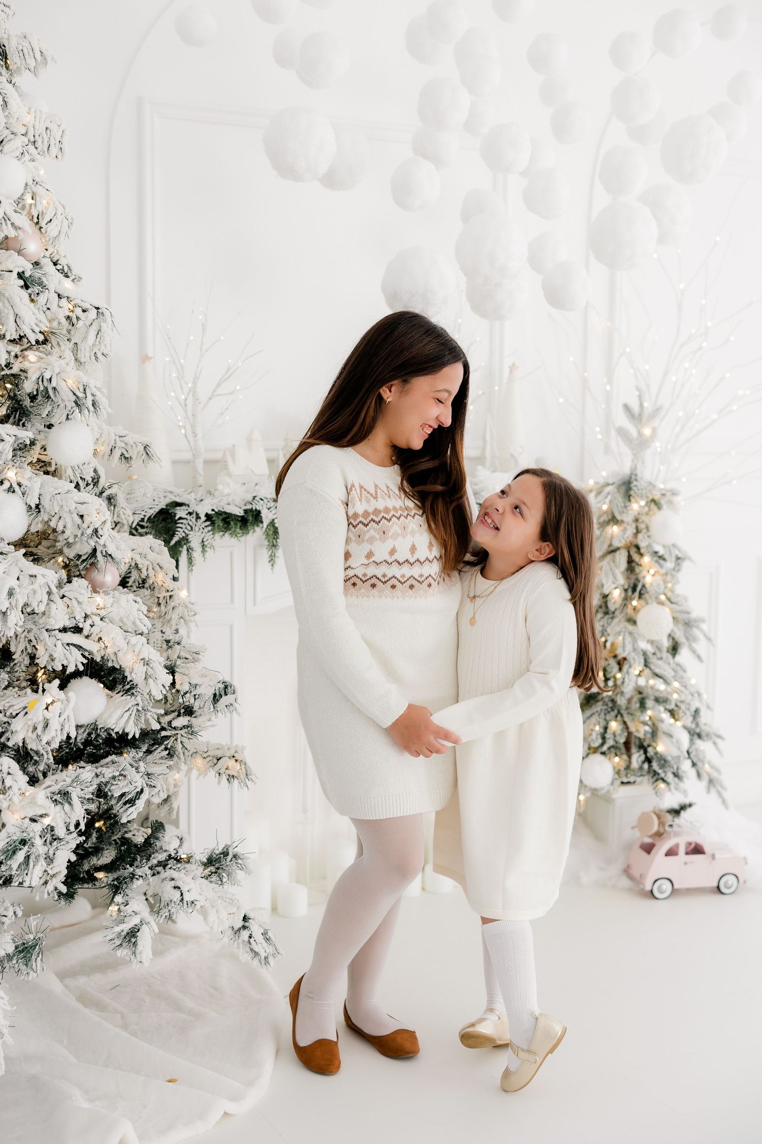 Mother and daughter wearing matching white sweaters pose in front of snowy Christmas decor.
