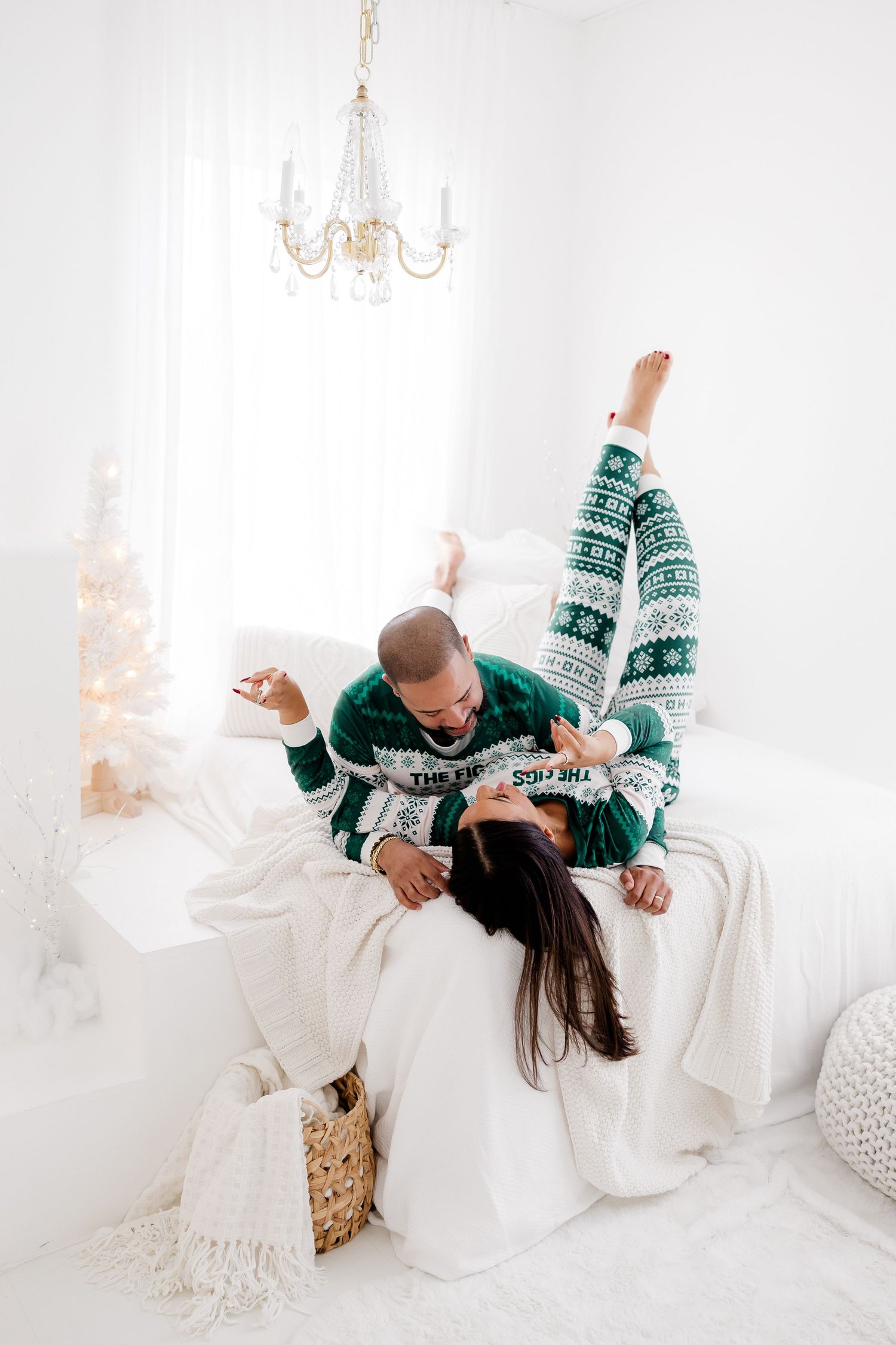 A playful couple in holiday sweaters lounges on a white couch beneath a crystal chandelier.