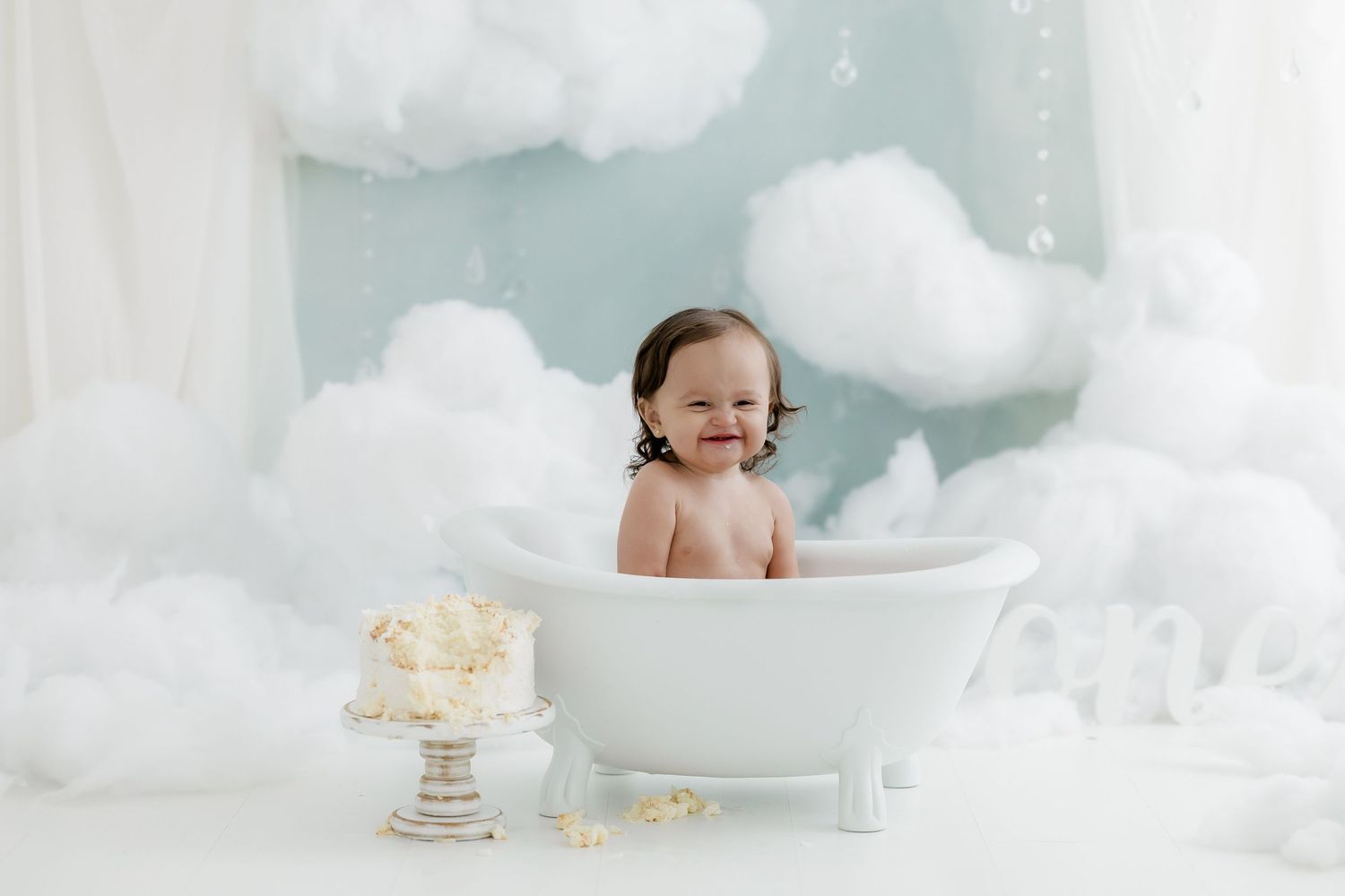 Happy baby sitting in a white bathtub surrounded by fluffy clouds with a smash cake nearby.