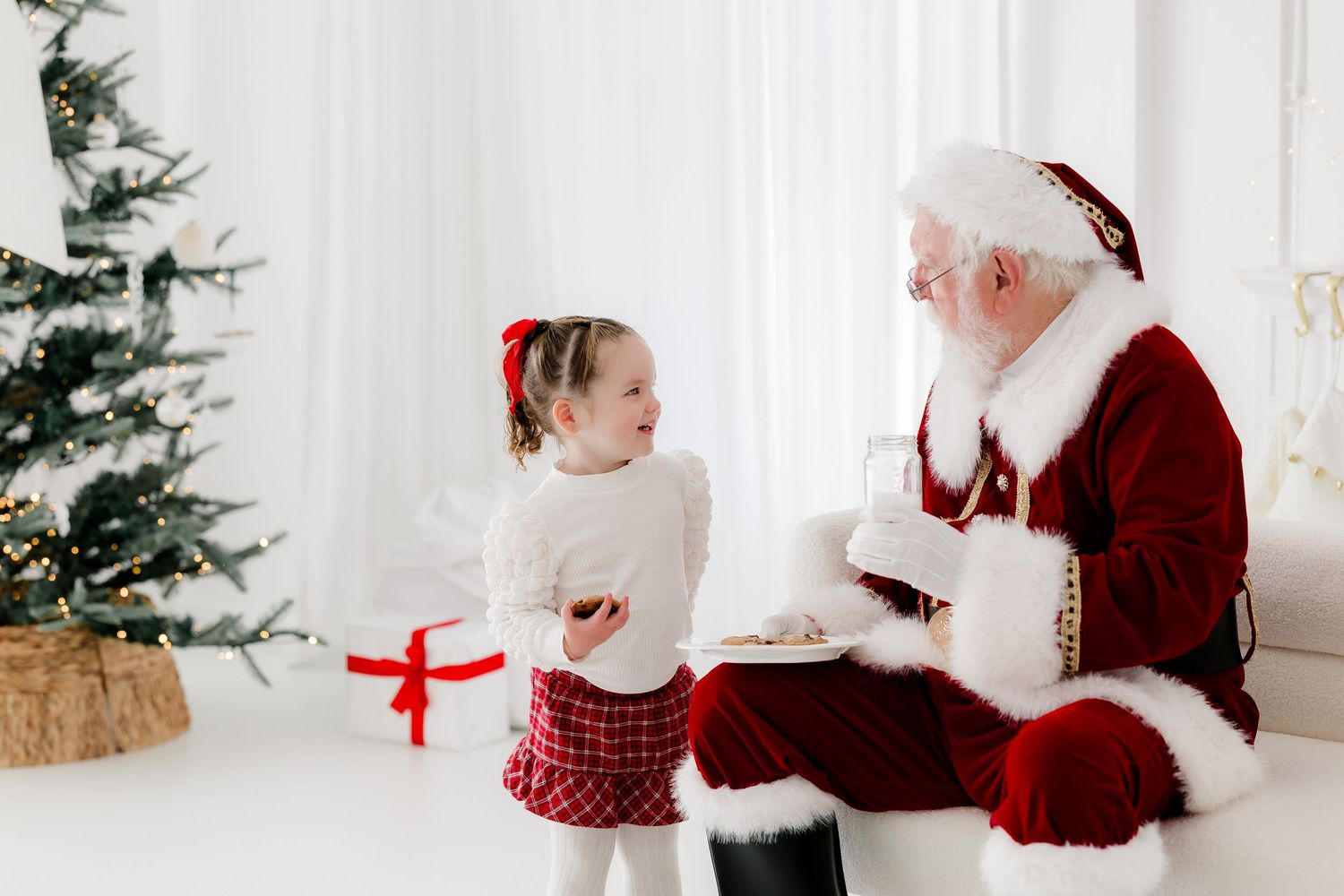 A young girl in a red plaid skirt talks to Santa Claus beside a decorated Christmas tree in a bright white room.