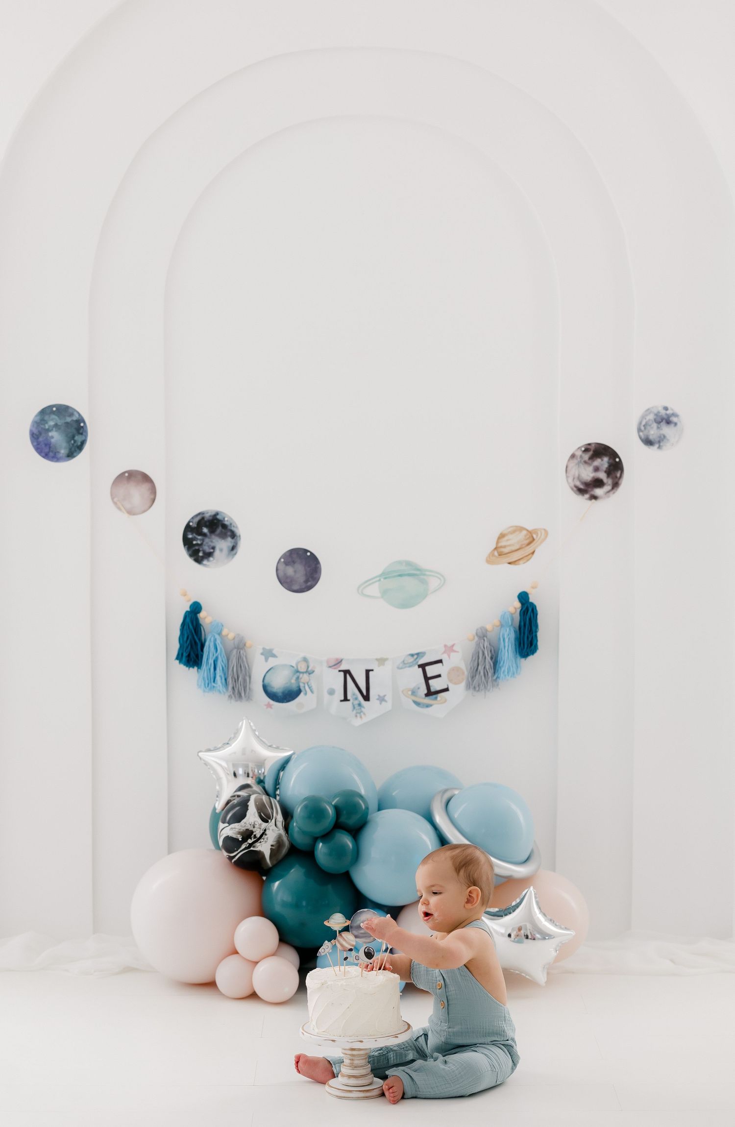 A space-themed first birthday cake smash setup with planets, balloons, and 'ONE' banner in blue and white colors.