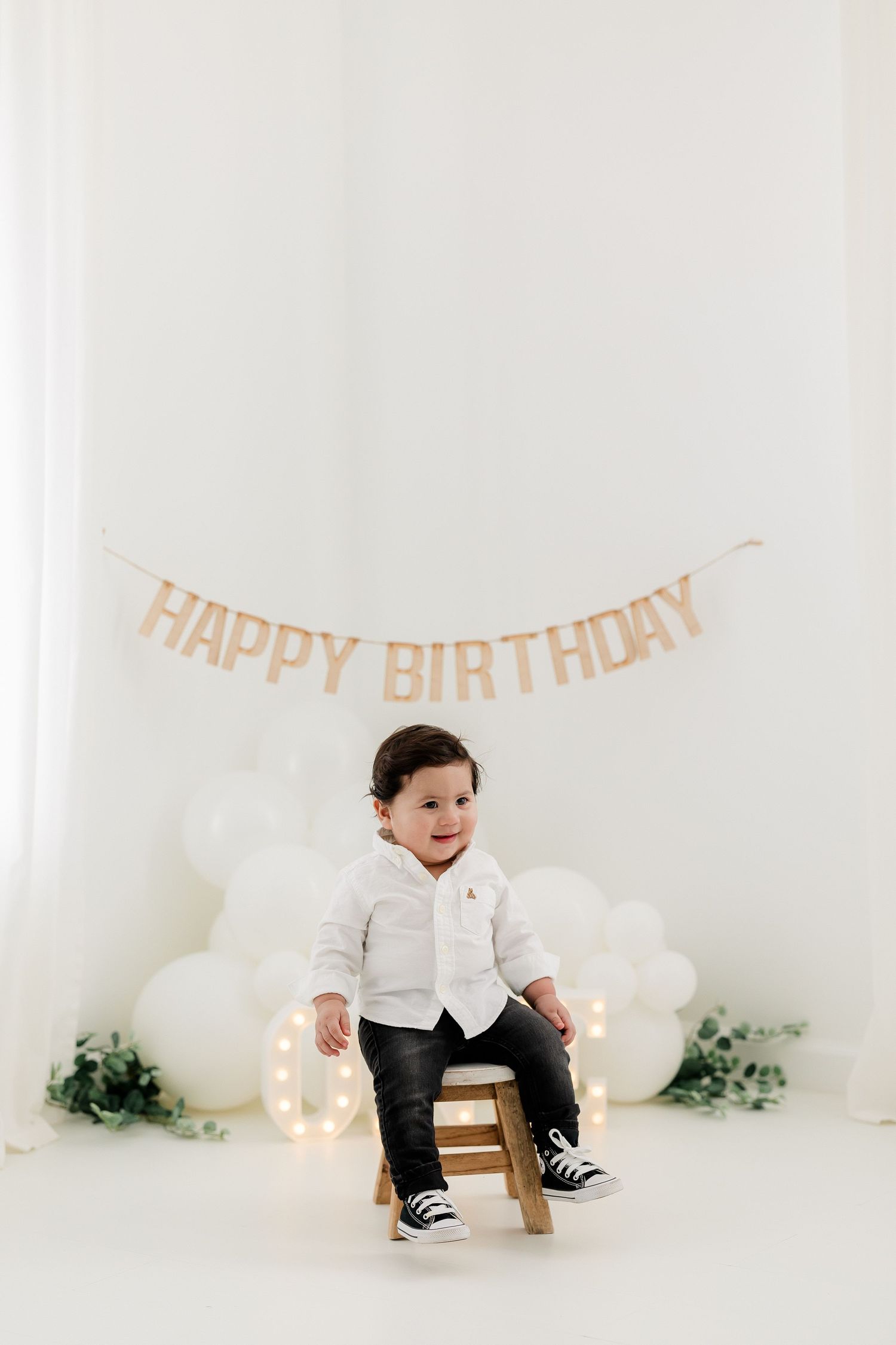 Minimalist first birthday setup with white balloons, wooden chair, and peach-colored 'Happy Birthday' banner against white backdrop.