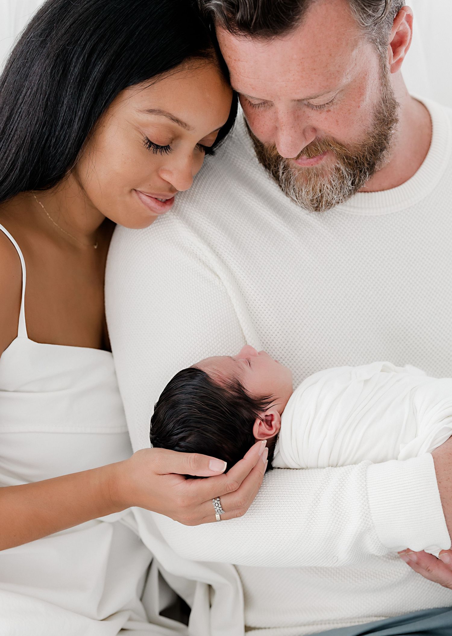 A tender moment between couple holding their newborn baby swaddled in white against light background.