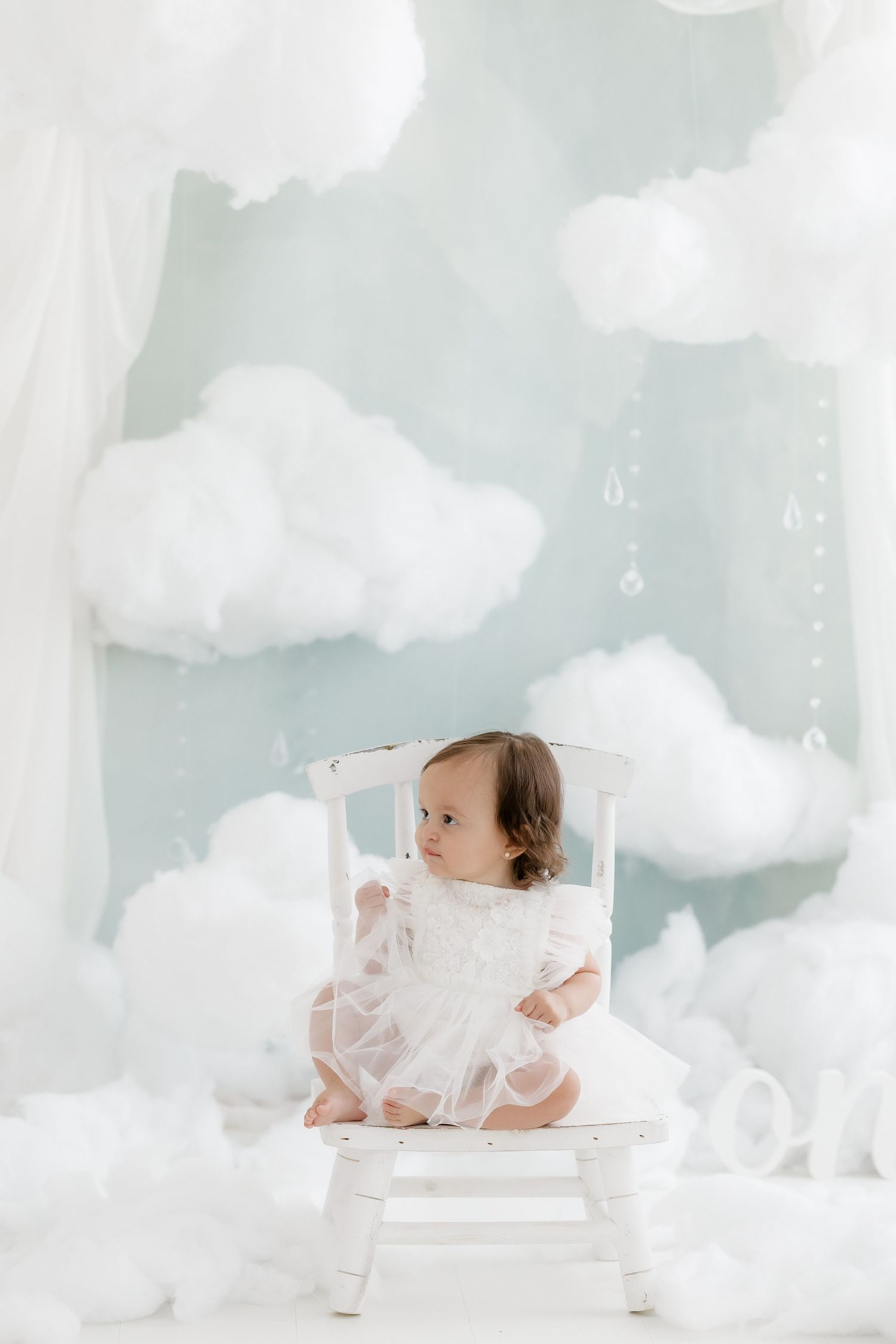 Baby girl in white dress sits on small white chair surrounded by fluffy white clouds in dreamy blue studio.