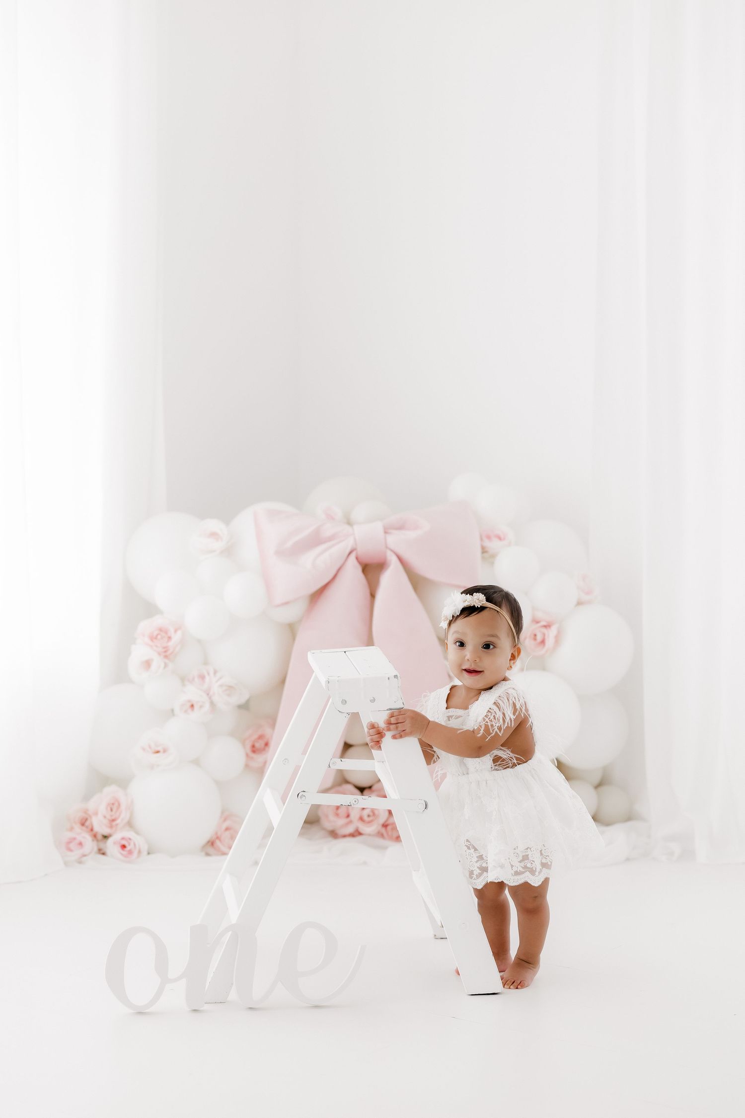 A baby in white dress sits on a pink and white themed set with floral decorations and balloons.