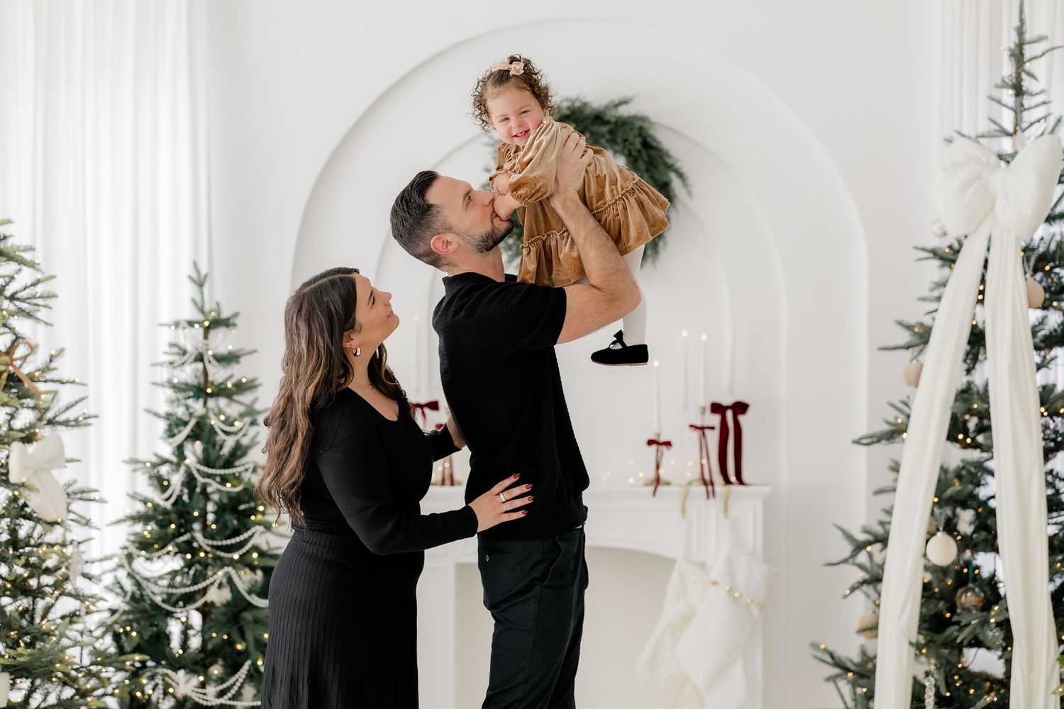 Family holiday photo with parents lifting toddler near decorated Christmas trees in bright white studio.