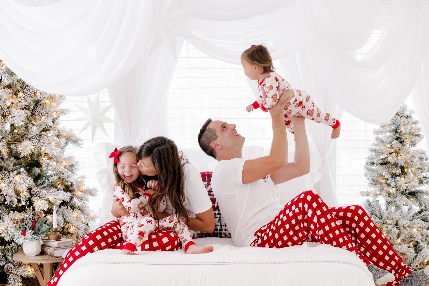 Family in matching Christmas pajamas on a bed surrounded by decorated trees, dad lifting toddler joyfully.