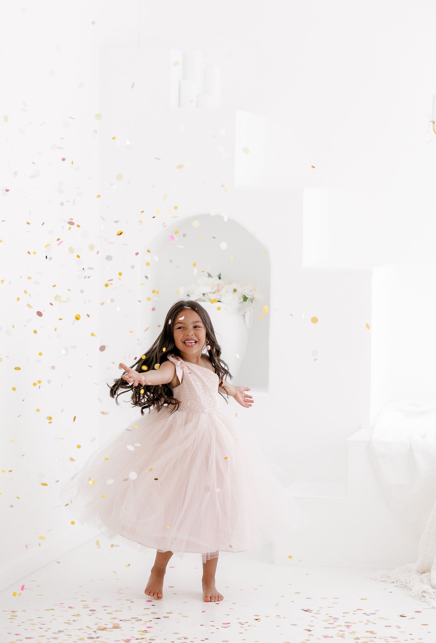 Young girl in a pink tulle dress twirls and dances among colorful confetti in a bright white studio setting.